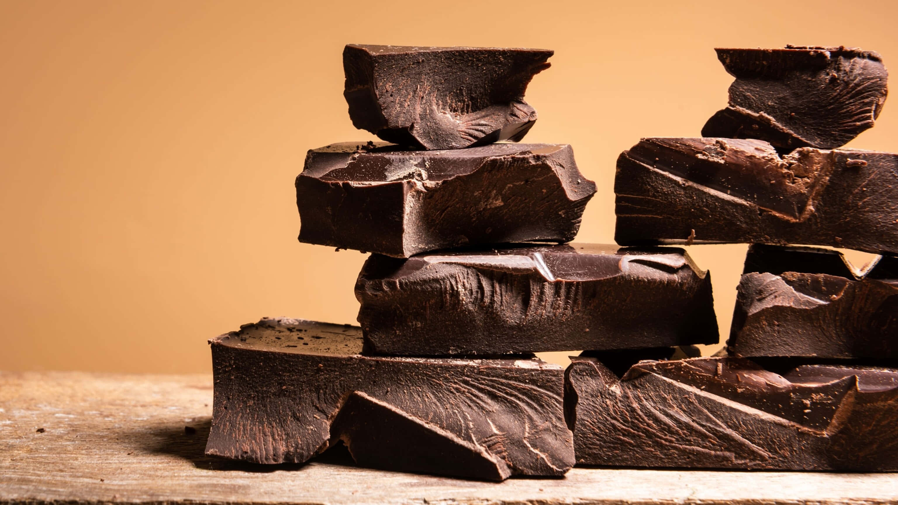 Dark Chocolate 桌布 壁紙 — 濃郁的黑朱古力塊不均勻地堆疊在質樸的木質表面上，背景是溫暖的米色。