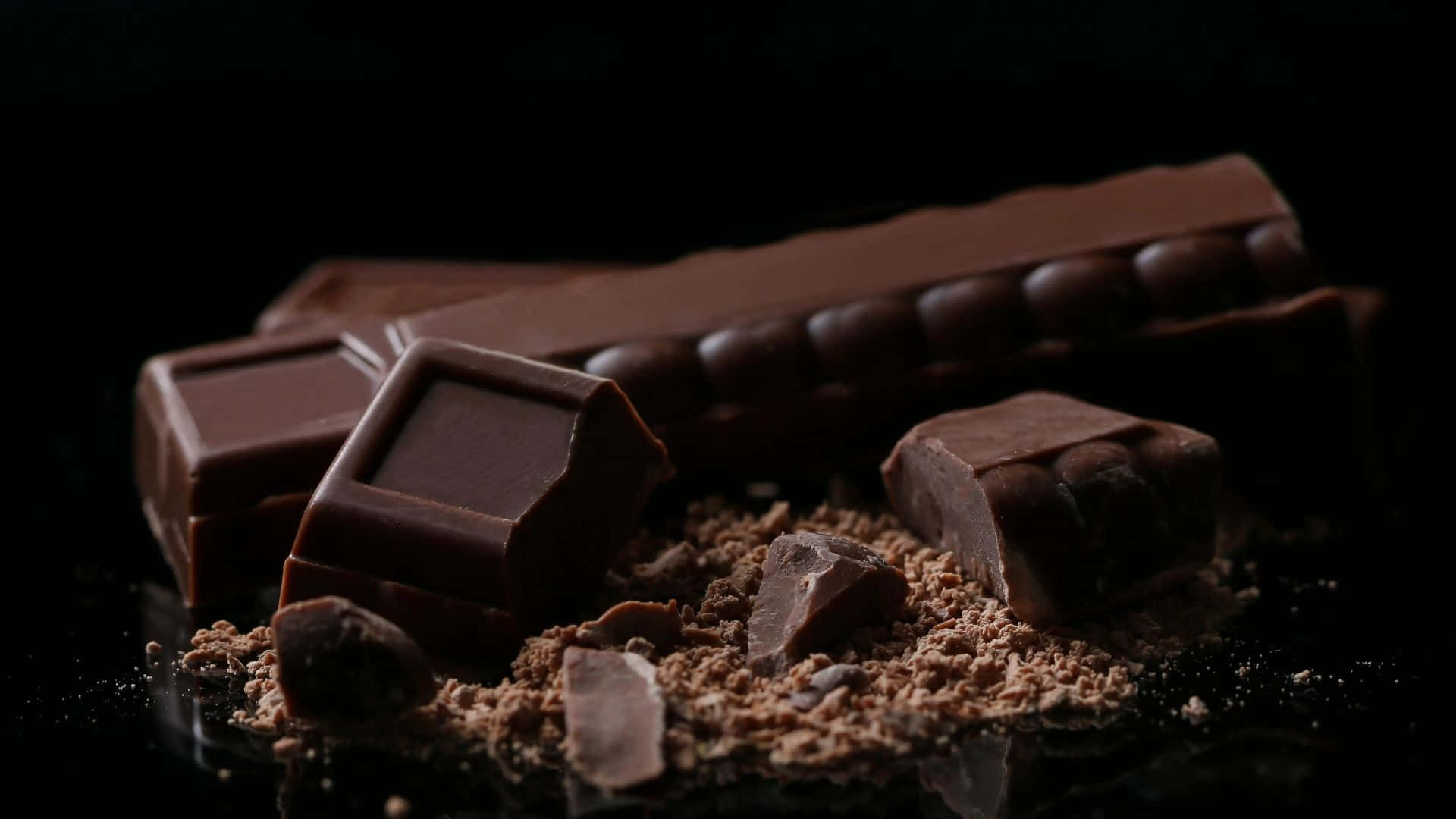 Dark Chocolate 桌布 壁紙 — 濃郁的黑朱古力條和碎塊，可可粉散落在光亮的黑色表面上，在戲劇性的燈光下。