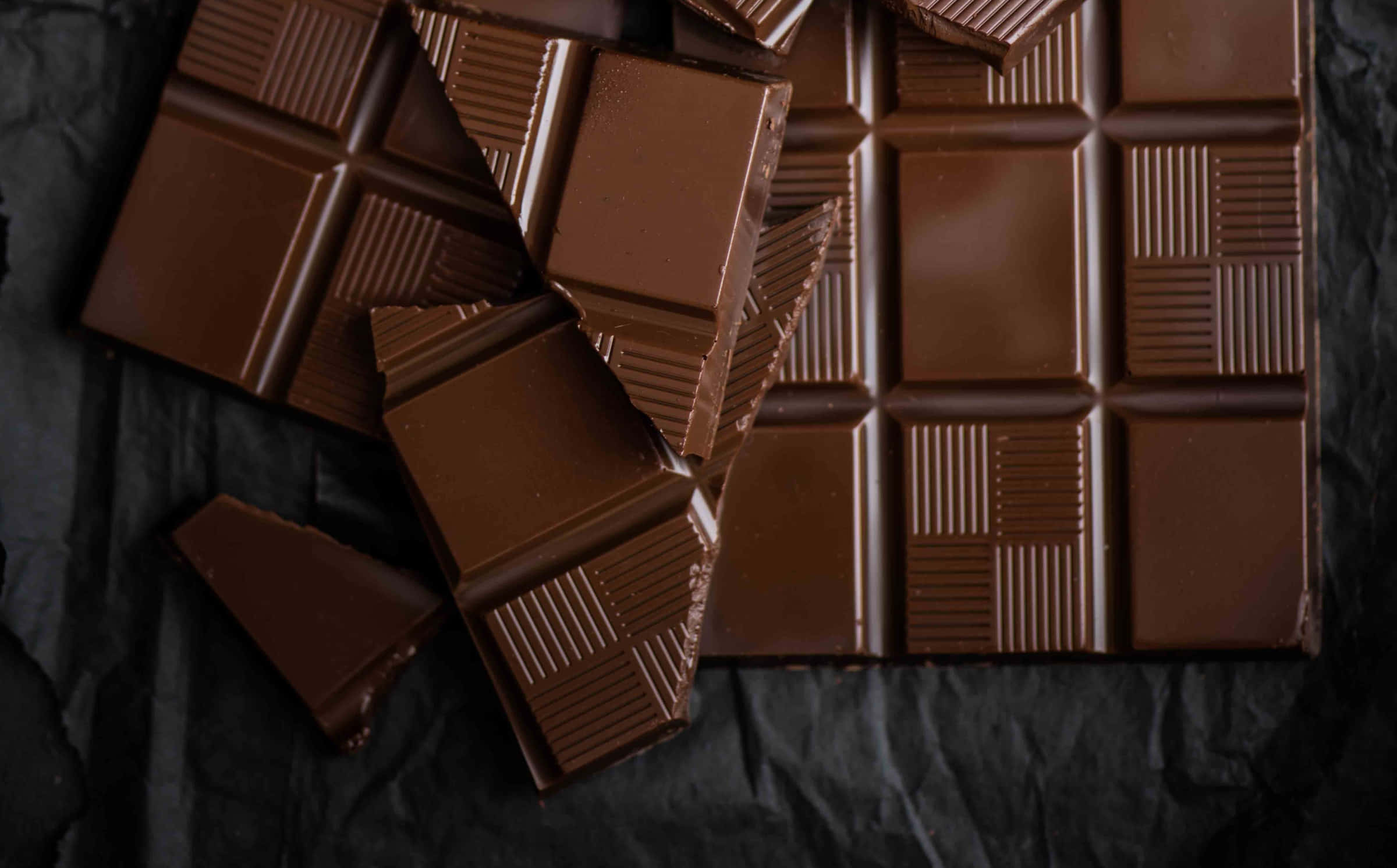 Brown Chocolate 桌布 壁紙 — 濃郁的黑朱古力條和碎塊放在皺巴巴的黑色紙上，帶有光澤質感和深棕色調。