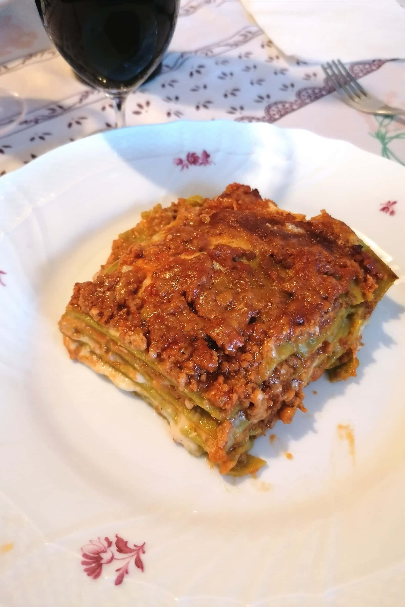 濃郁肉醬波隆那千層麵 壁紙 — 一塊金黃色的千層麵，配上濃郁的番茄醬和融化的芝士，放在有紅色花卉圖案的白色碟子上。