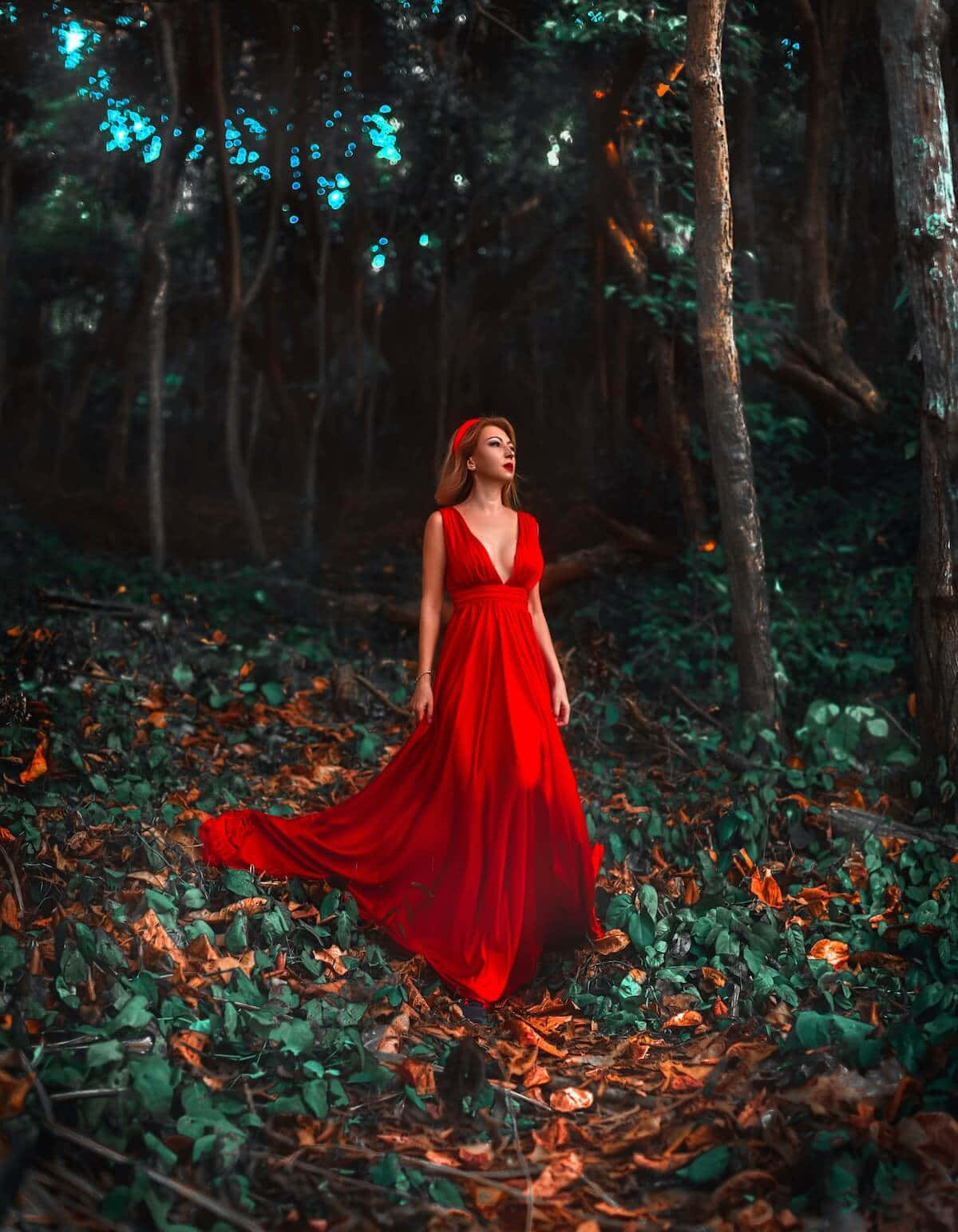 濃郁鮮豔的紅色審美，為任何場景注入活力 壁紙 — 一位身穿飄逸紅色長裙的女性站在茂密的森林地面上，地面覆蓋著落葉，在斑駁的藍色和綠色光線下。