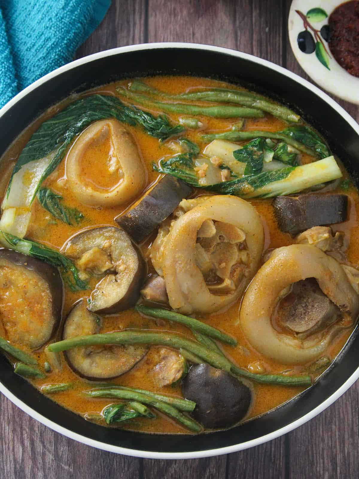 濃醬菲律賓燉菜 Kare-Kare 壁紙 — 一個木桌上放著一碗冒著熱氣的濃郁橙色咖喱，配有魷魚圈、茄子和青豆。