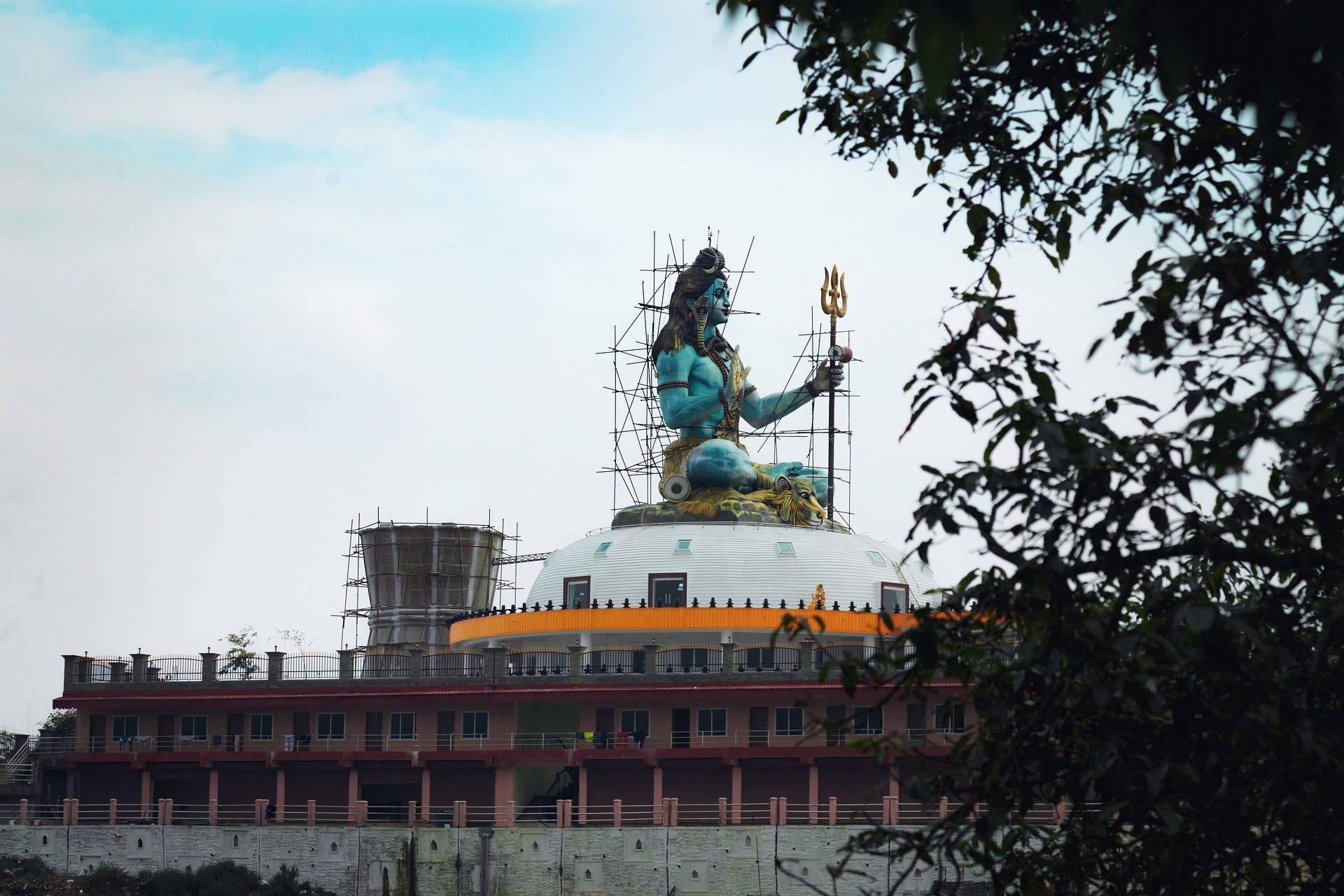 被樹枝環繞的寺廟圓頂上，一座正在建造中的大型藍色濕婆神像。