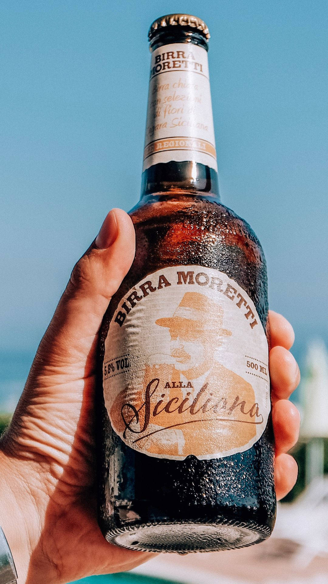 濕潤的義大利啤酒Bira Moretti 壁紙 — 一隻手抓住一瓶冰凍的Birra Moretti Alla Siciliana，背景是明亮的藍天，隱約可見海洋。