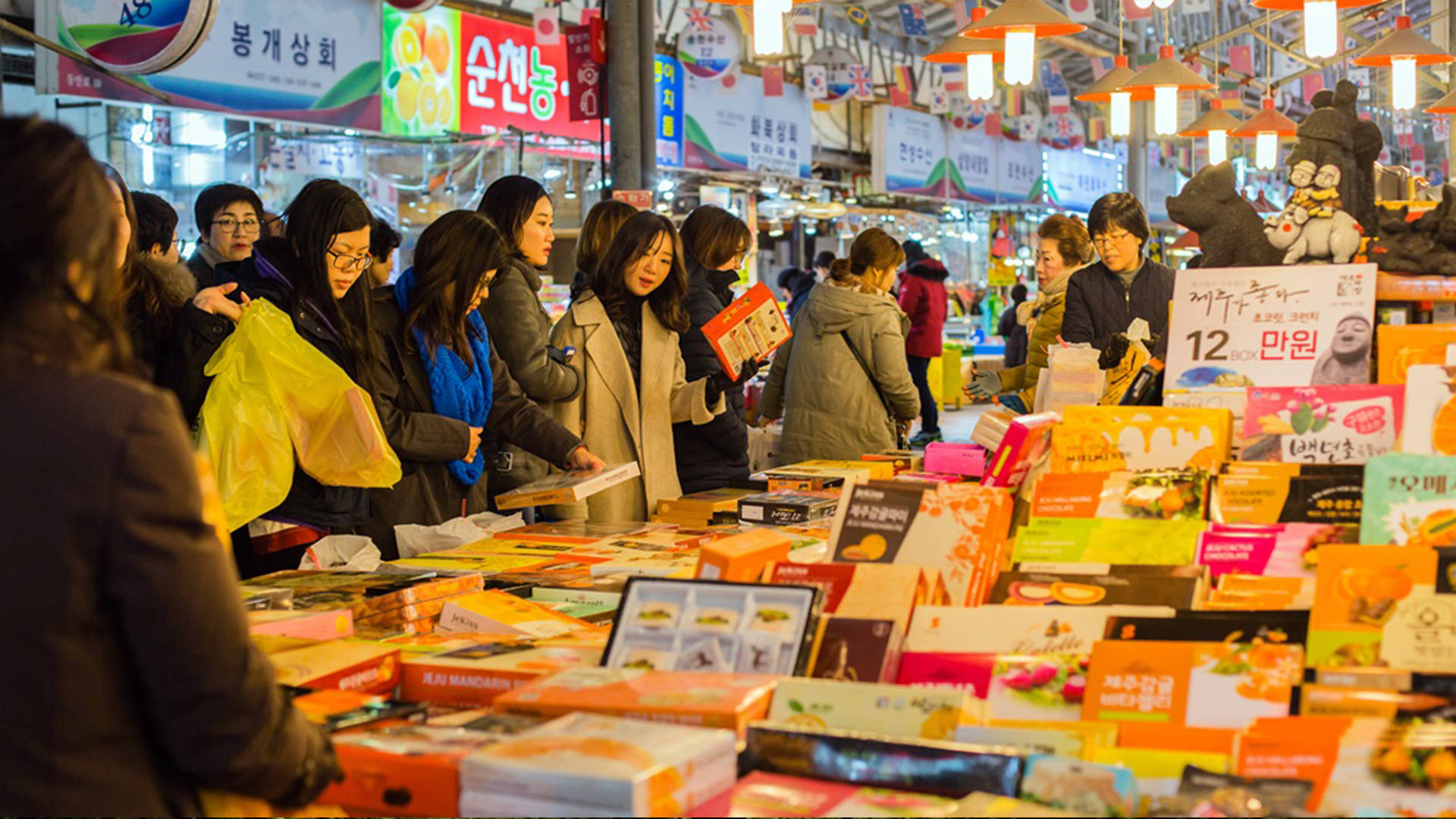 濟州島東門市場的迷人風貌 壁紙 — 擁擠的夜市攤位堆滿了色彩繽紛的包裝商品，購物者在明亮的吊燈下瀏覽。