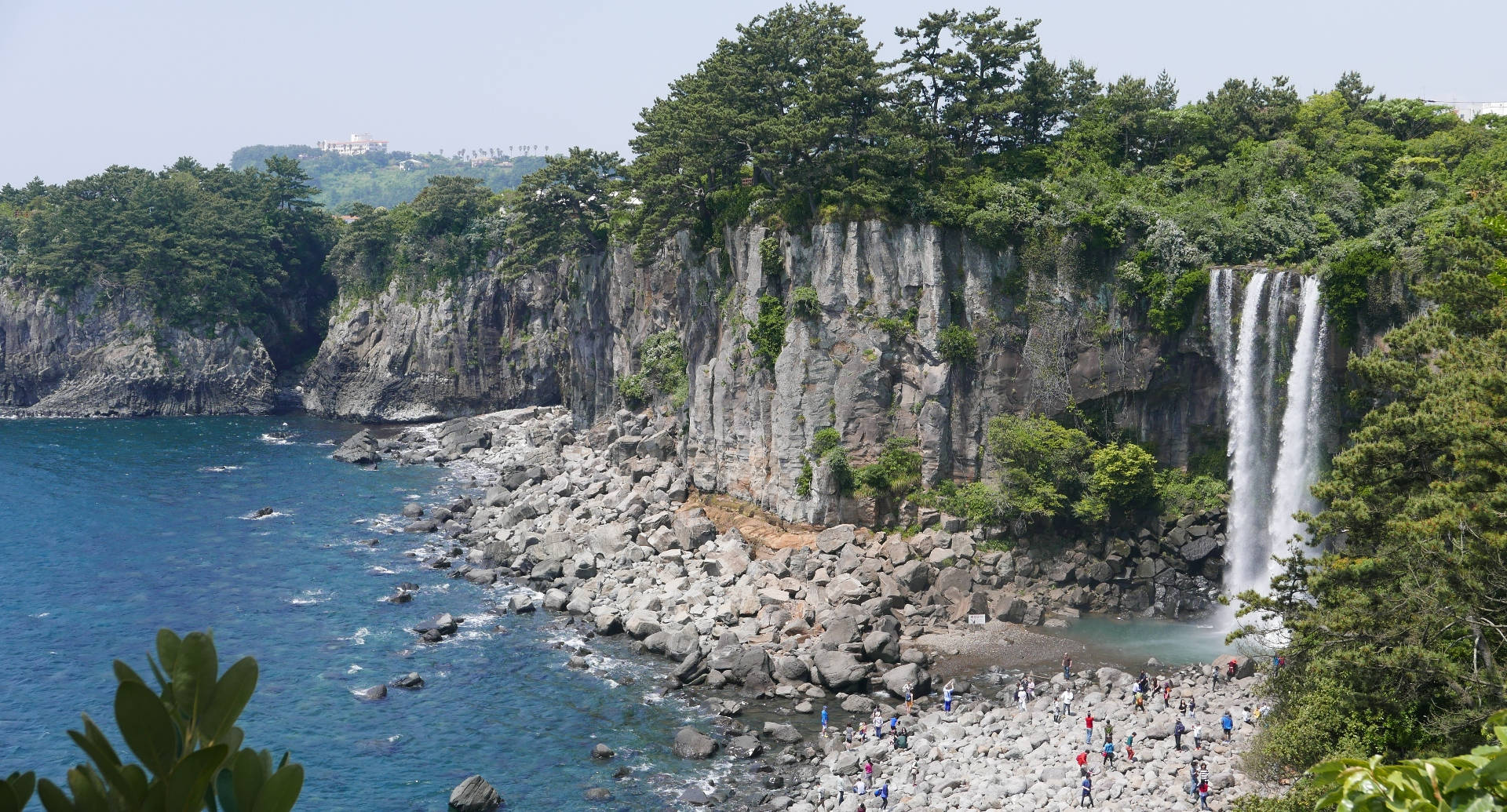 濟州島正房瀑布 壁紙 — 一個宏偉的海岸瀑布從崎嶇的懸崖傾瀉而下，流入碧綠色的海洋，遊客聚集在岩石海岸上。