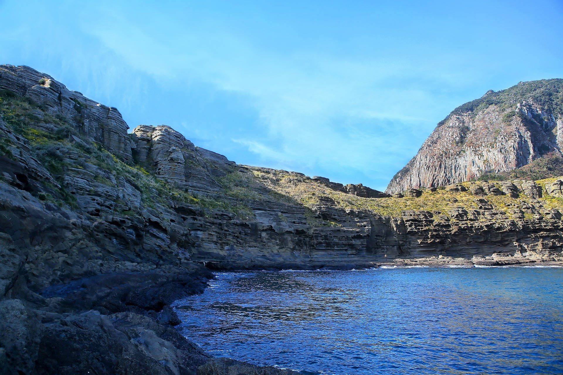 濟州島龍梅里海岸 壁紙 — 一個寧靜的海灣，深藍色的海水隱藏在崎嶇的層狀懸崖之間，上方是清澈的藍天。