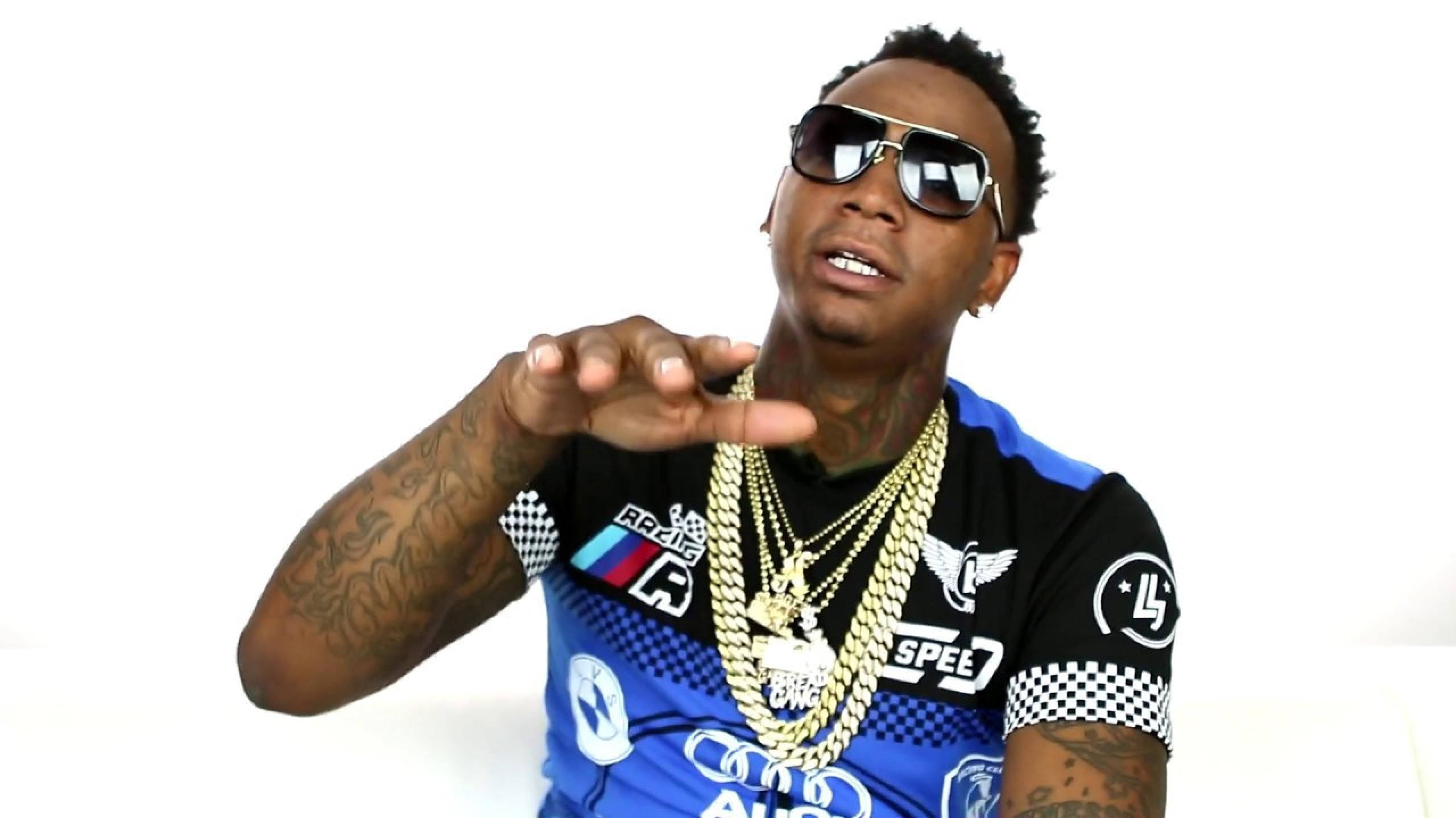 瀟灑 Moneybagg Yo 壁紙 — 一位時尚男士身穿藍色賽車服和金色鏈條，在純白背景前自信地擺姿勢。