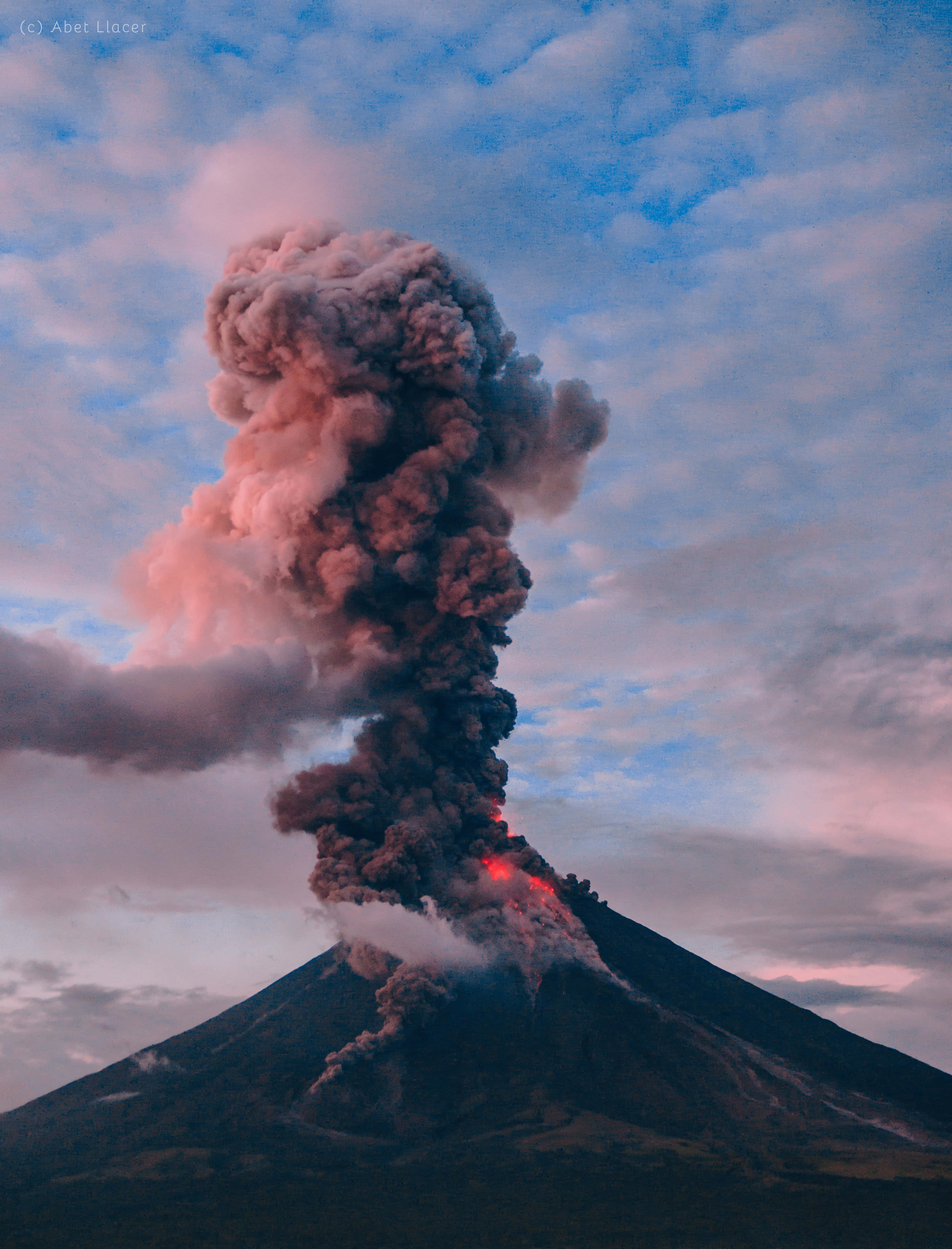 火山口上方燦爛橙色日出 壁紙 — 一座火山在黃昏時猛烈噴發，將發光的熔岩和濃煙噴向粉紅色和藍色的天空。
