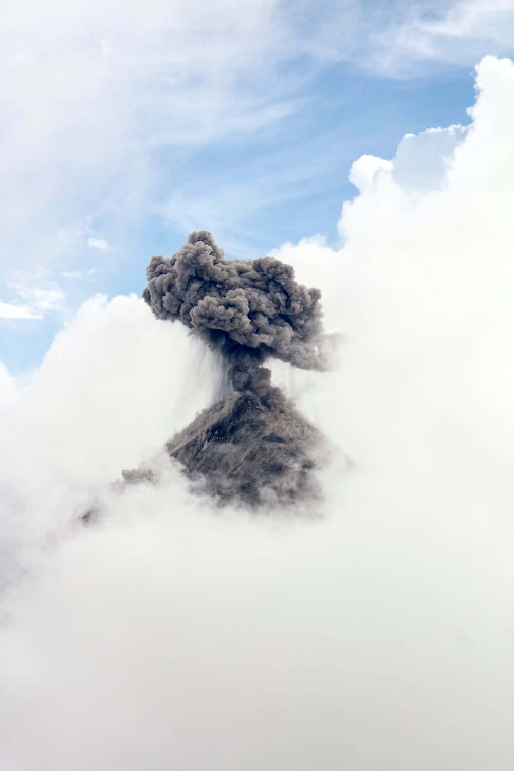 火山爆發的壯麗日落景色 壁紙 — 火山爆發將深色火山灰噴向被 fluffy 白色雲朵環繞的明亮藍天高空。