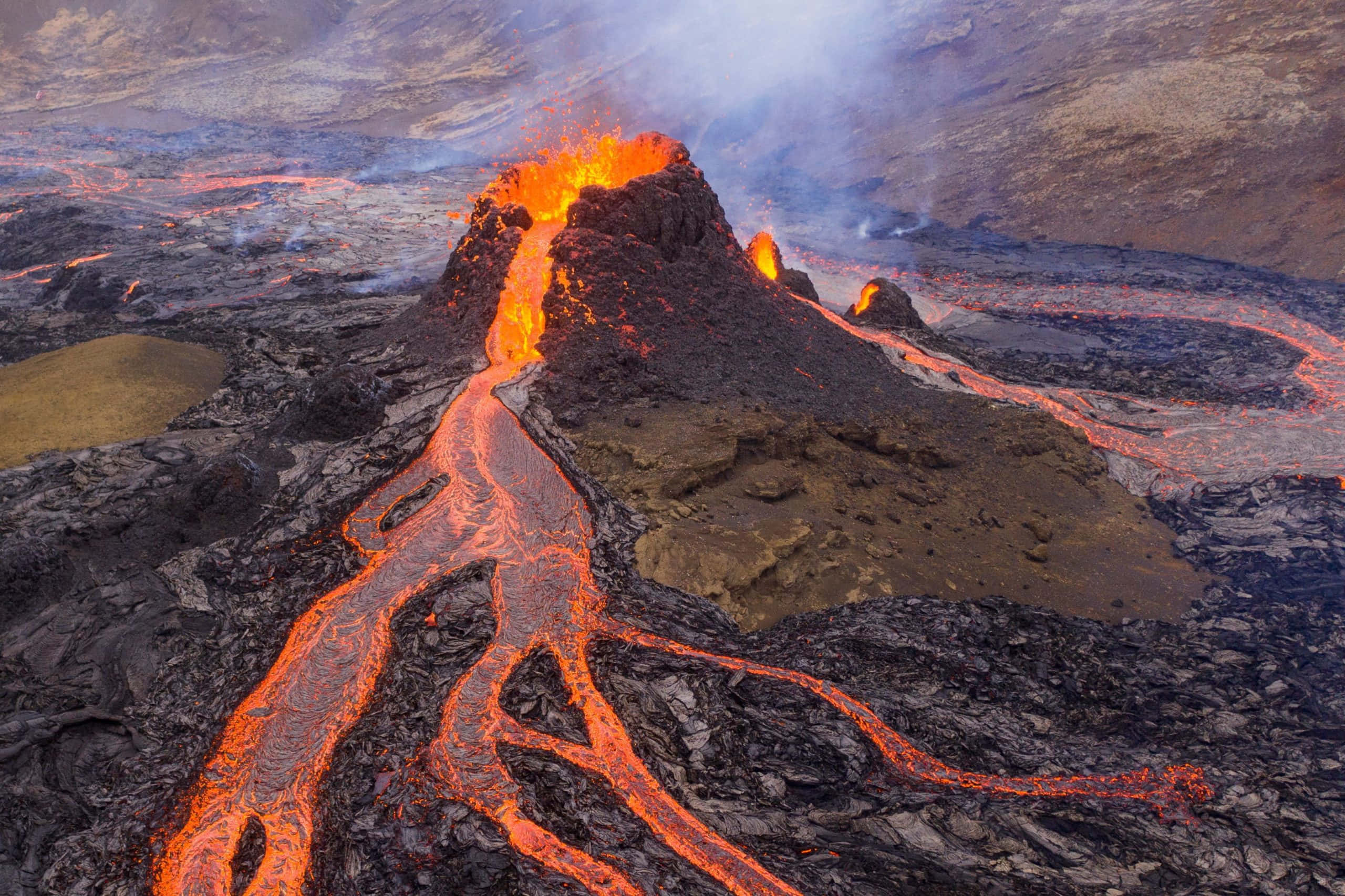 火山的空中壯麗視角 壁紙 — 火山爆發在昏暗的天空下，將熾熱的橙色熔岩噴灑在崎嶇的黑色岩層上。