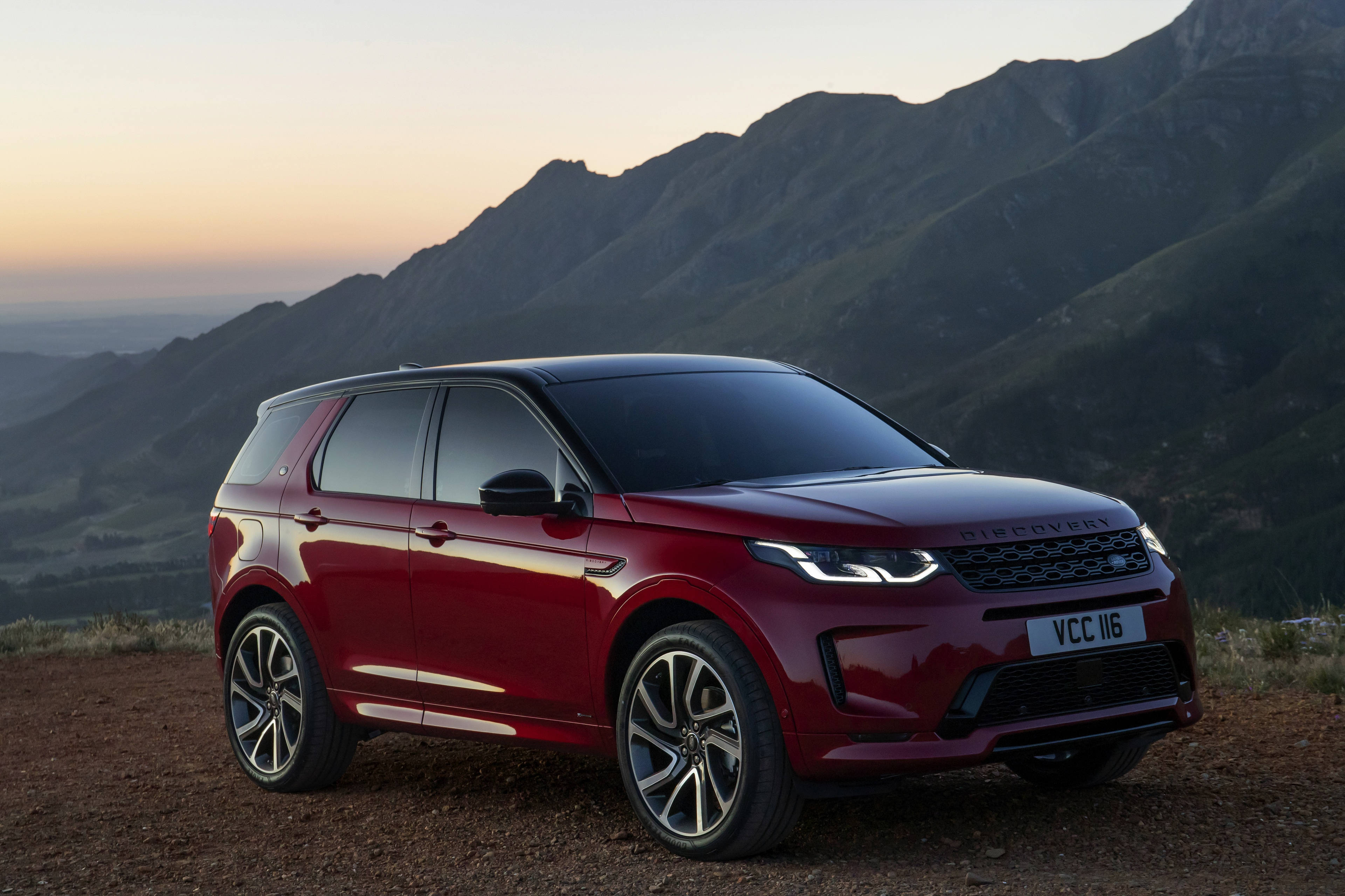 火烈鳥紅色發現運動版路虎 iPhone 主題壁紙 壁紙 — 一架亮紅色嘅 Land Rover Discovery 泊喺一個 dirt 觀景點，背景係起伏嘅綠色山脈同柔和嘅日落天空。