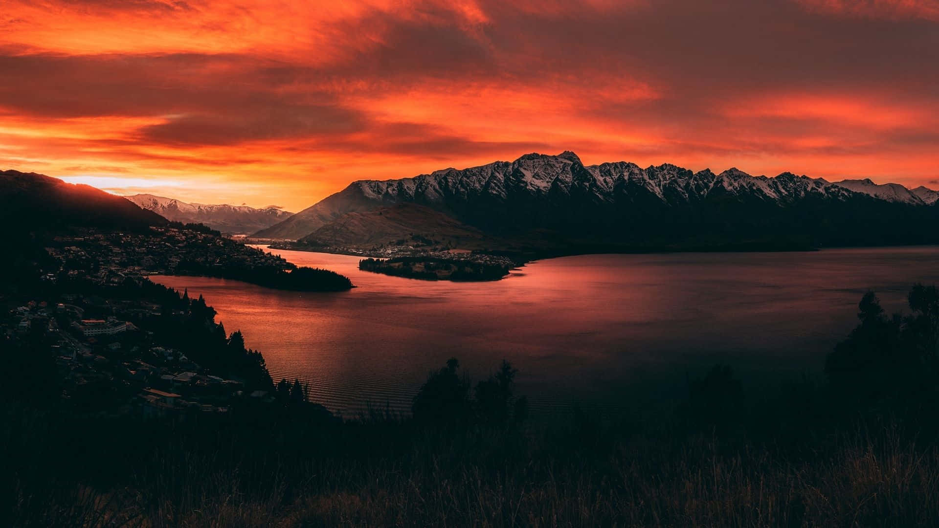 Orange Sunset 桌布 壁紙 — 火熱的橙色夕陽染紅了天空，並倒映在平靜的湖面上，周圍是白雪覆蓋的山脈和一個小鎮。