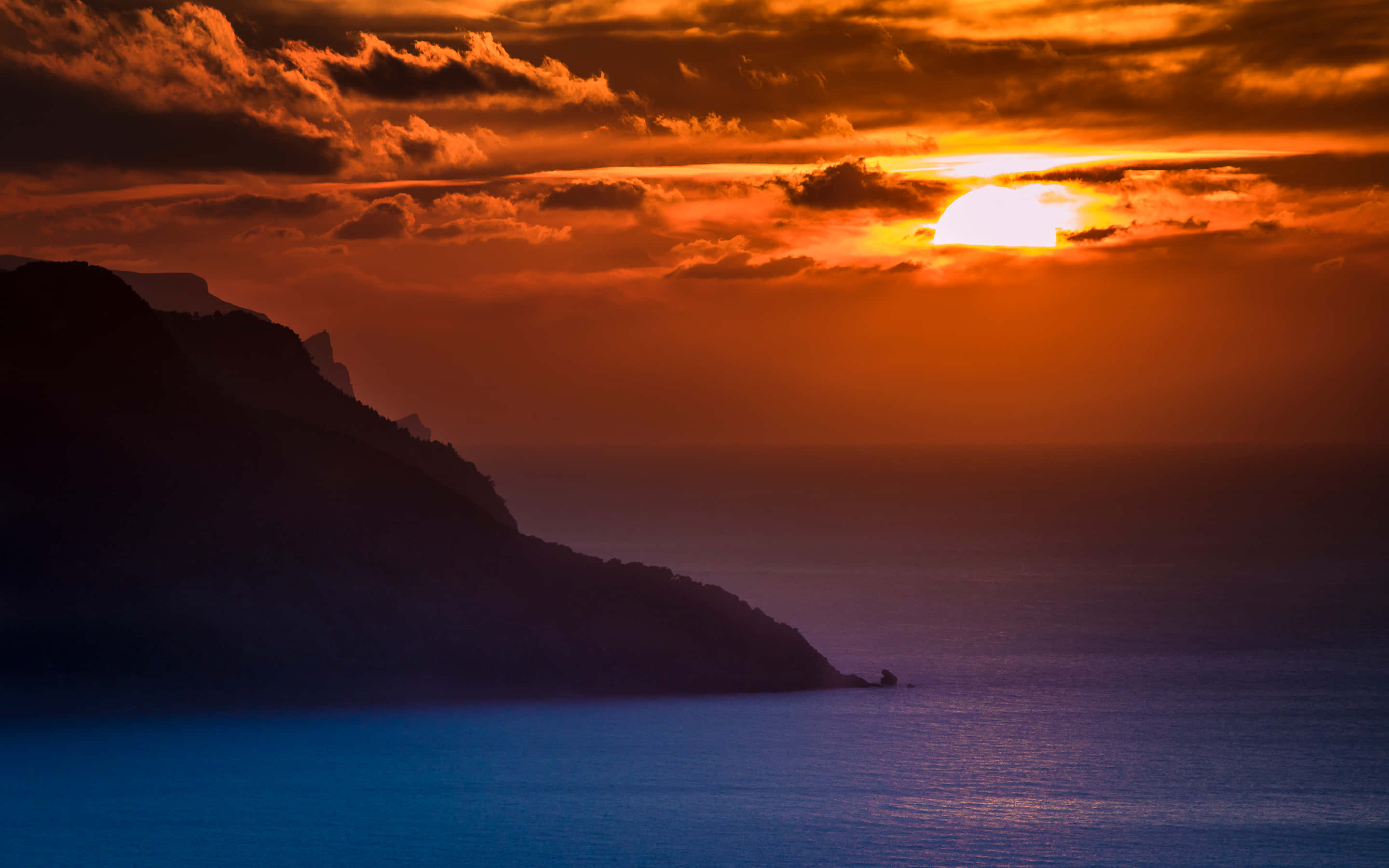 Sunset Ocean 桌布 壁紙 — 火熱的橙色太陽在雲層後落下，在黑暗的懸崖和深藍色的海洋上投射出溫暖的光芒。