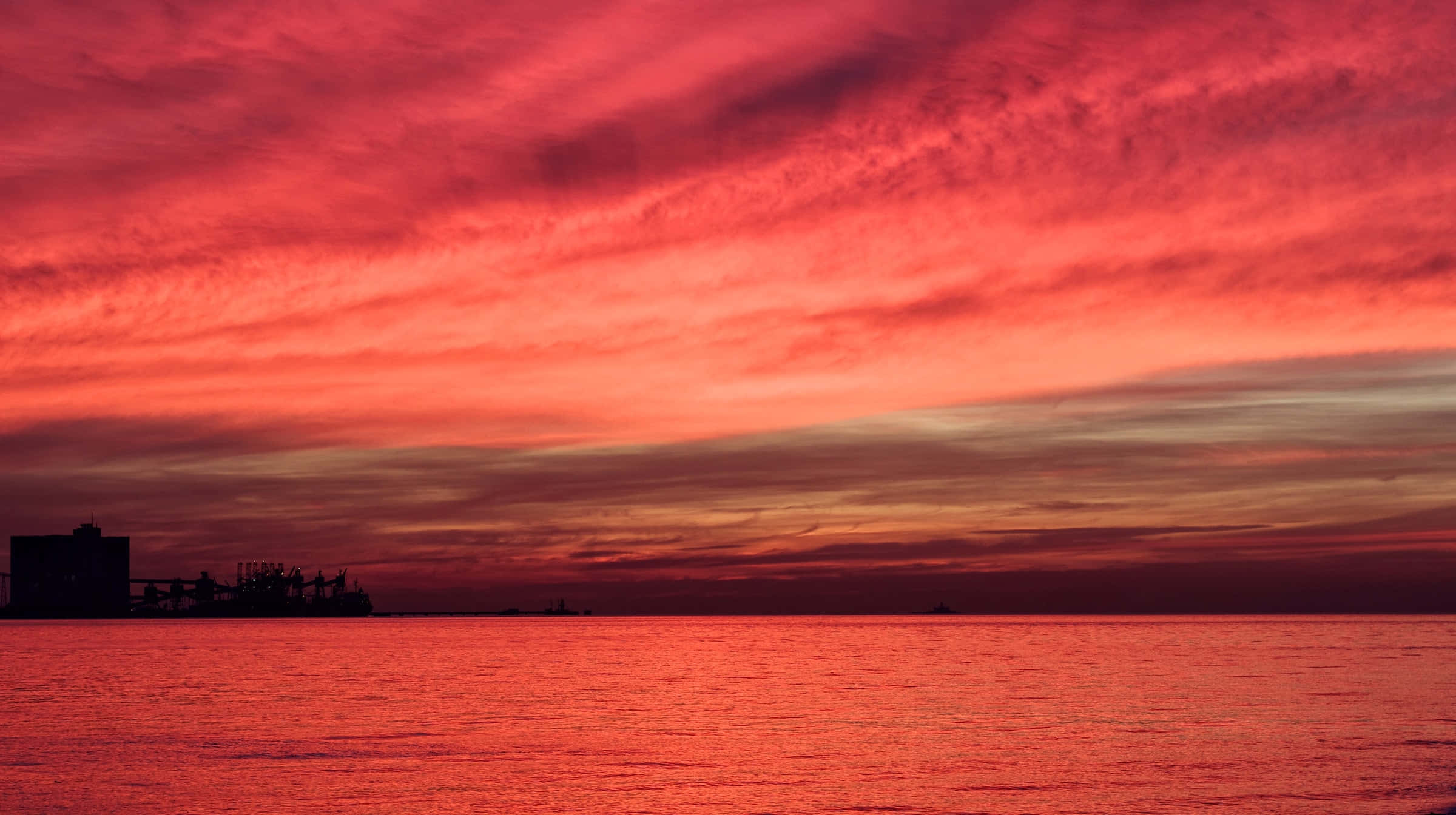 Red Sunset 桌布 壁紙 — 火紅的夕陽染紅了天空和海洋，海岸建築的黑色剪影矗立在地平線上。