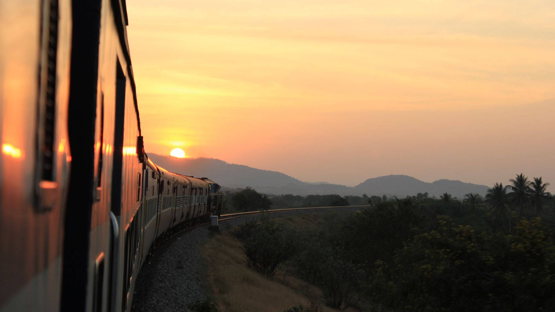 火車夕陽山景之旅 壁紙 — 一列火車在日落時滑過，在朦朧的天空和遠山背景下，在其側面投下金色的光芒。