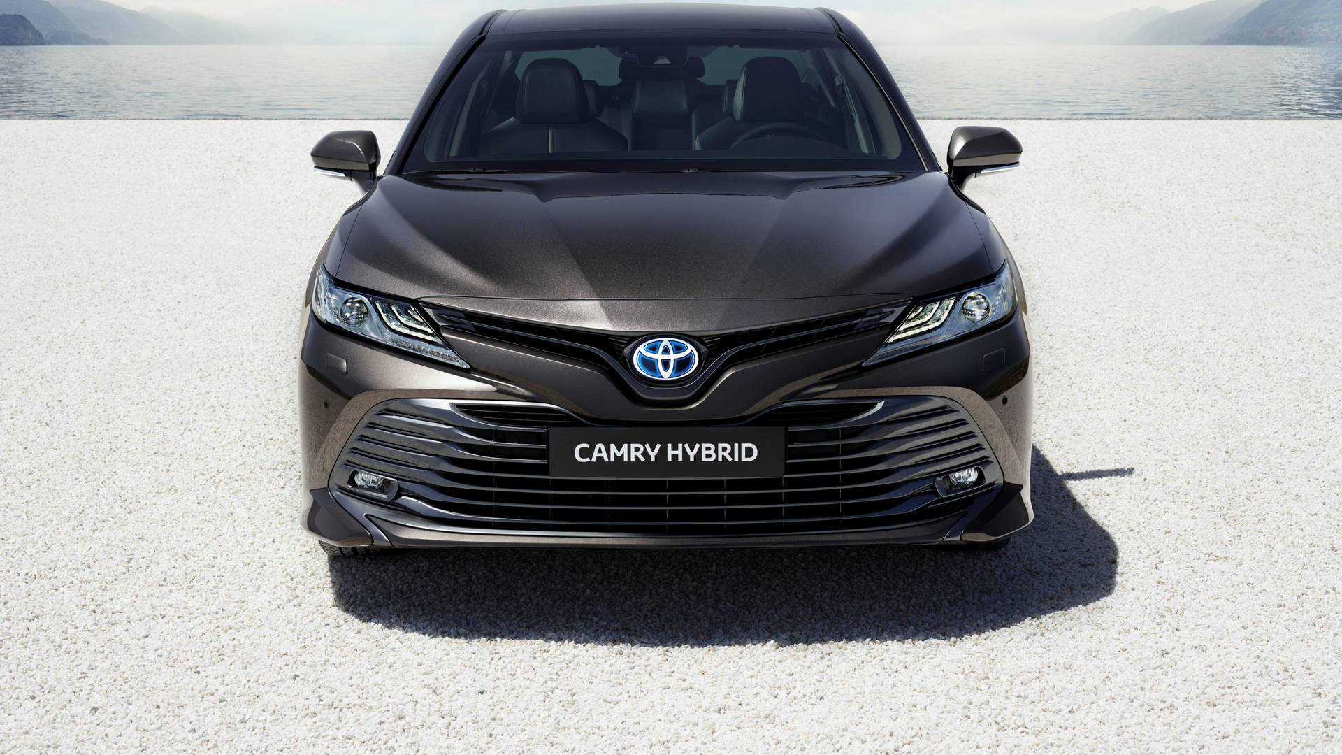 深灰色豐田Camry Hybrid的前視圖，在礫石上靠近水邊，背景有山脈。