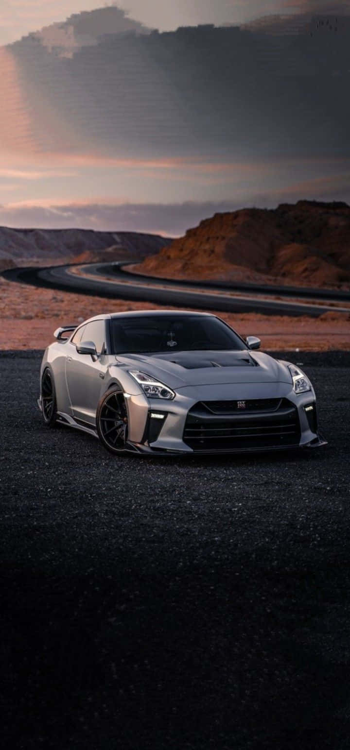 灰色 Nissan R35 GT-R 立體肖像圖 壁紙 — 一架時尚的銀色跑車停泊在黑暗的碎石路上，天空呈現戲劇性的黃昏色彩，帶有橙色和紫色的雲彩。
