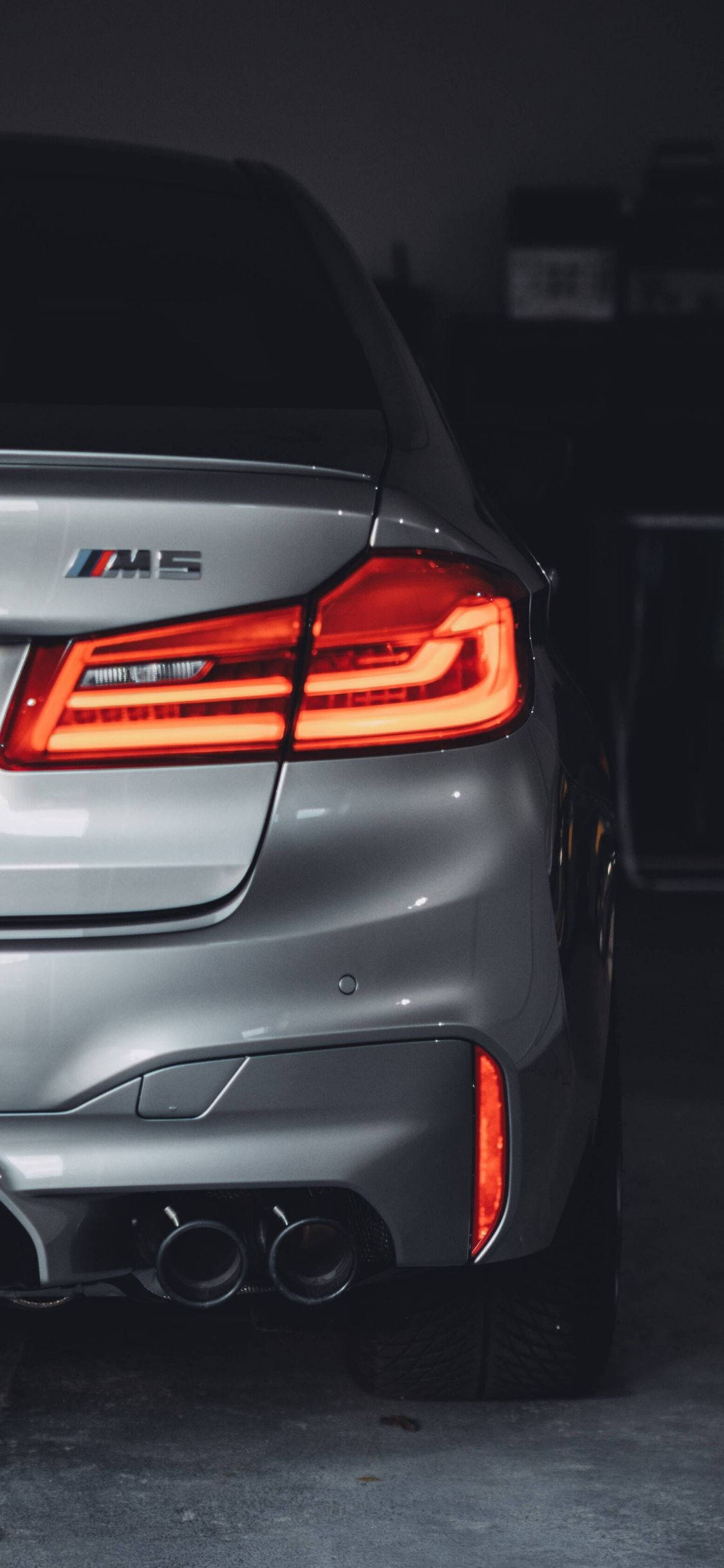 灰色BMW M5汽車iPhone壁紙 壁紙 — 特寫銀色寶馬M5在昏暗車庫中發光的紅色尾燈和雙排氣管。