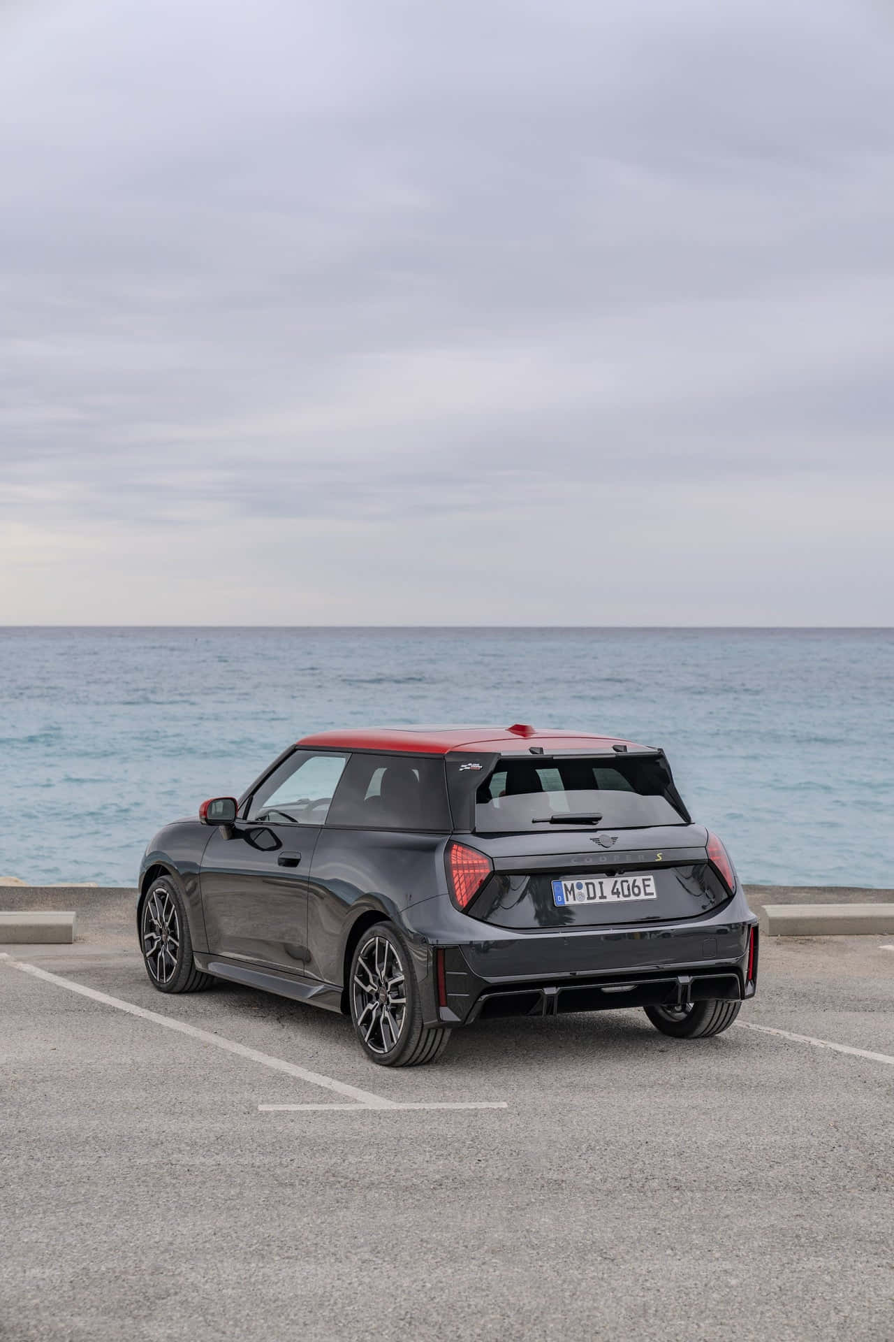 灰色Mini Cooper SE停泊於海邊 壁紙 — 一輛時尚的黑色MINI，紅色車頂，停泊在俯瞰平靜藍色海洋的沿海停車場，天空多雲。