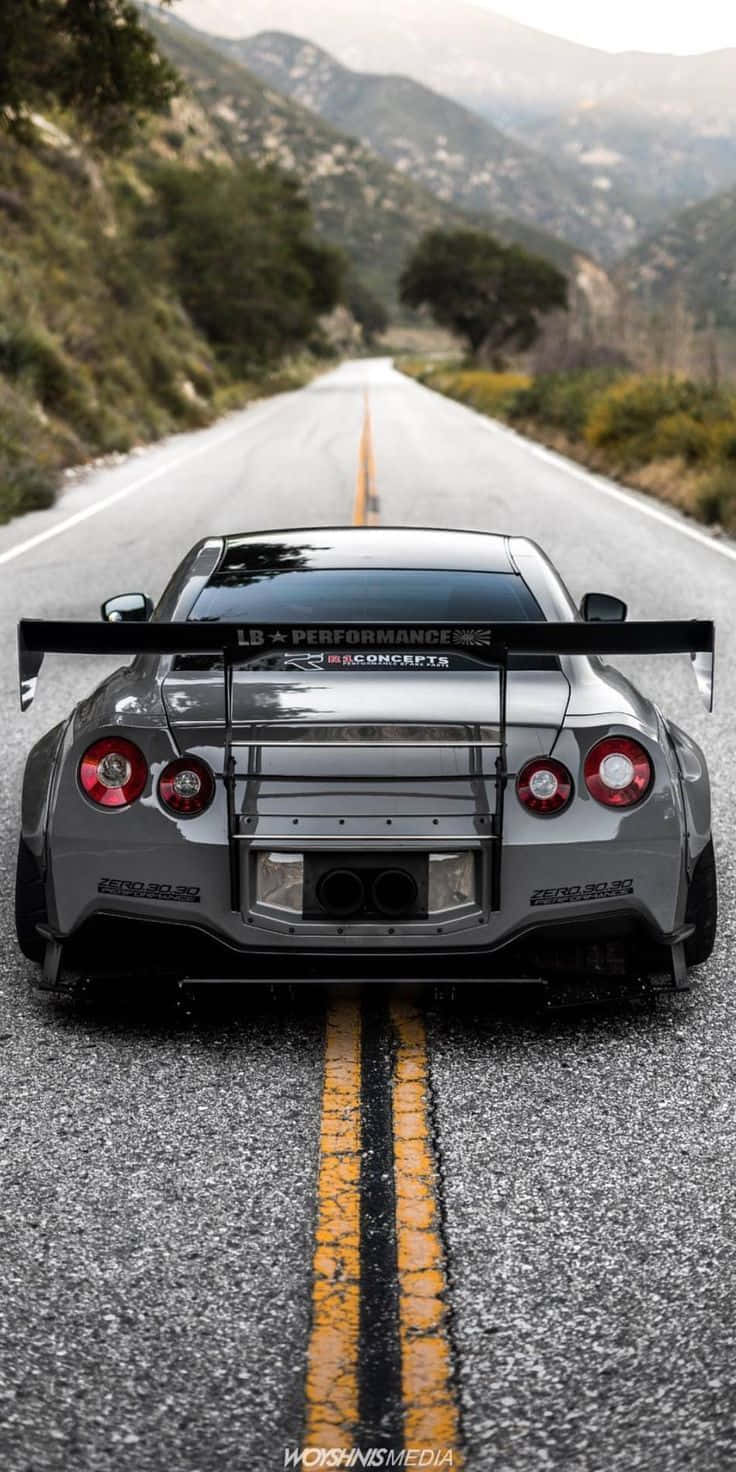 灰色Nissan R35 GT-R道路後視圖 壁紙 — 一架銀色改裝跑車配備大型尾翼，停泊在蜿蜒的山路上，路面有黃色中心線。