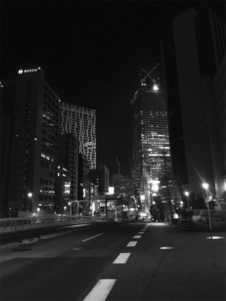 灰色生活的美感 壁紙 — 一條黑白夜景城市街道，兩旁是高聳的摩天大樓，窗戶亮著燈，街燈投下長長的影子。