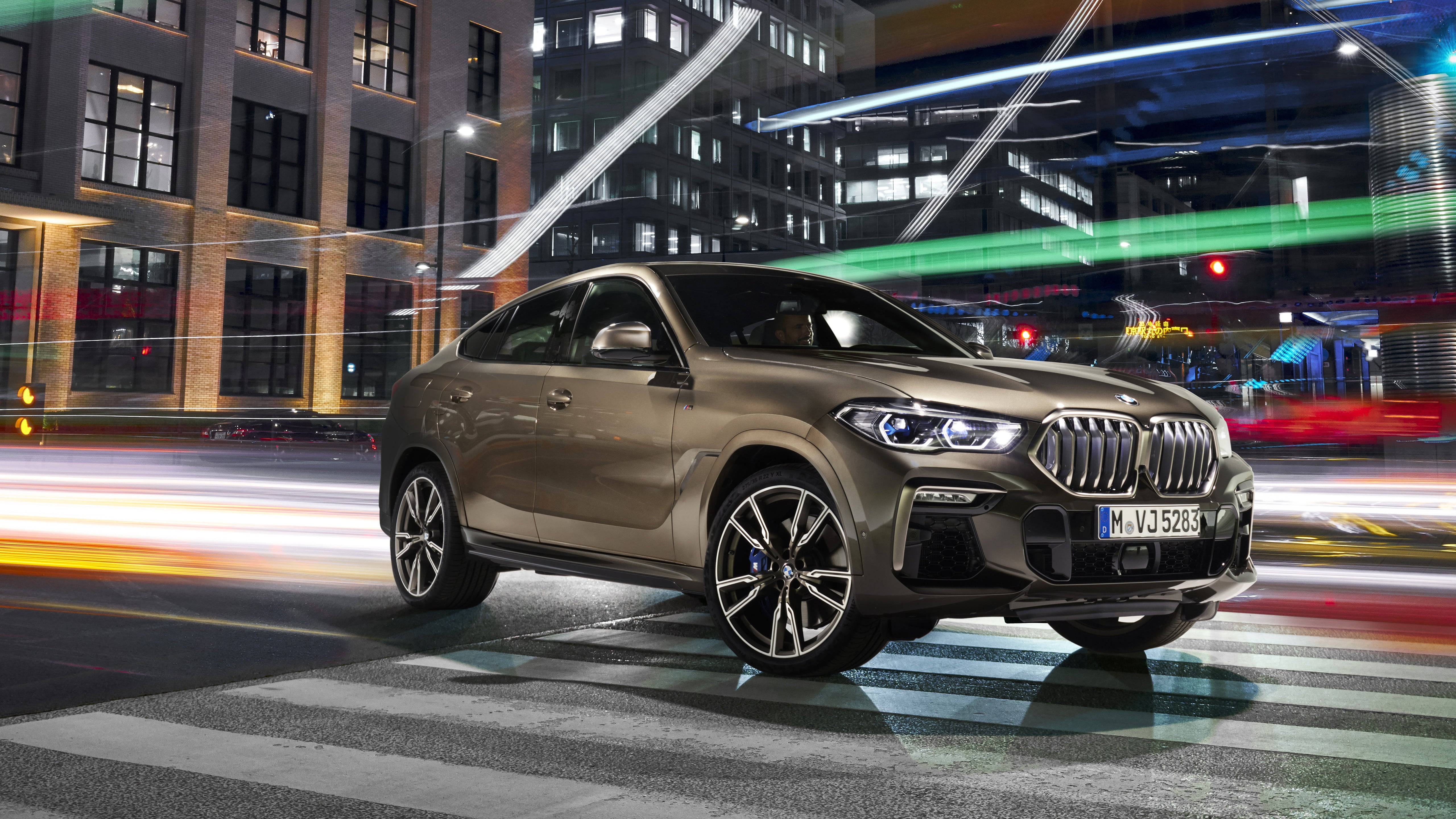 灰銀色BMW X6 M行駛於高速公路 壁紙 — 一輛時尚的金色BMW X6 SUV在夜晚穿過充滿活力的城市街道，身後是霓虹燈光和模糊的車流。