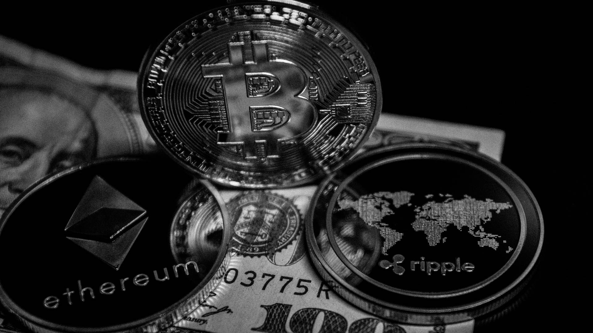 灰階以太幣硬幣 壁紙 — 單色特寫比特幣、以太坊和瑞波幣加密貨幣硬幣堆疊在美元鈔票上，背景為深色。