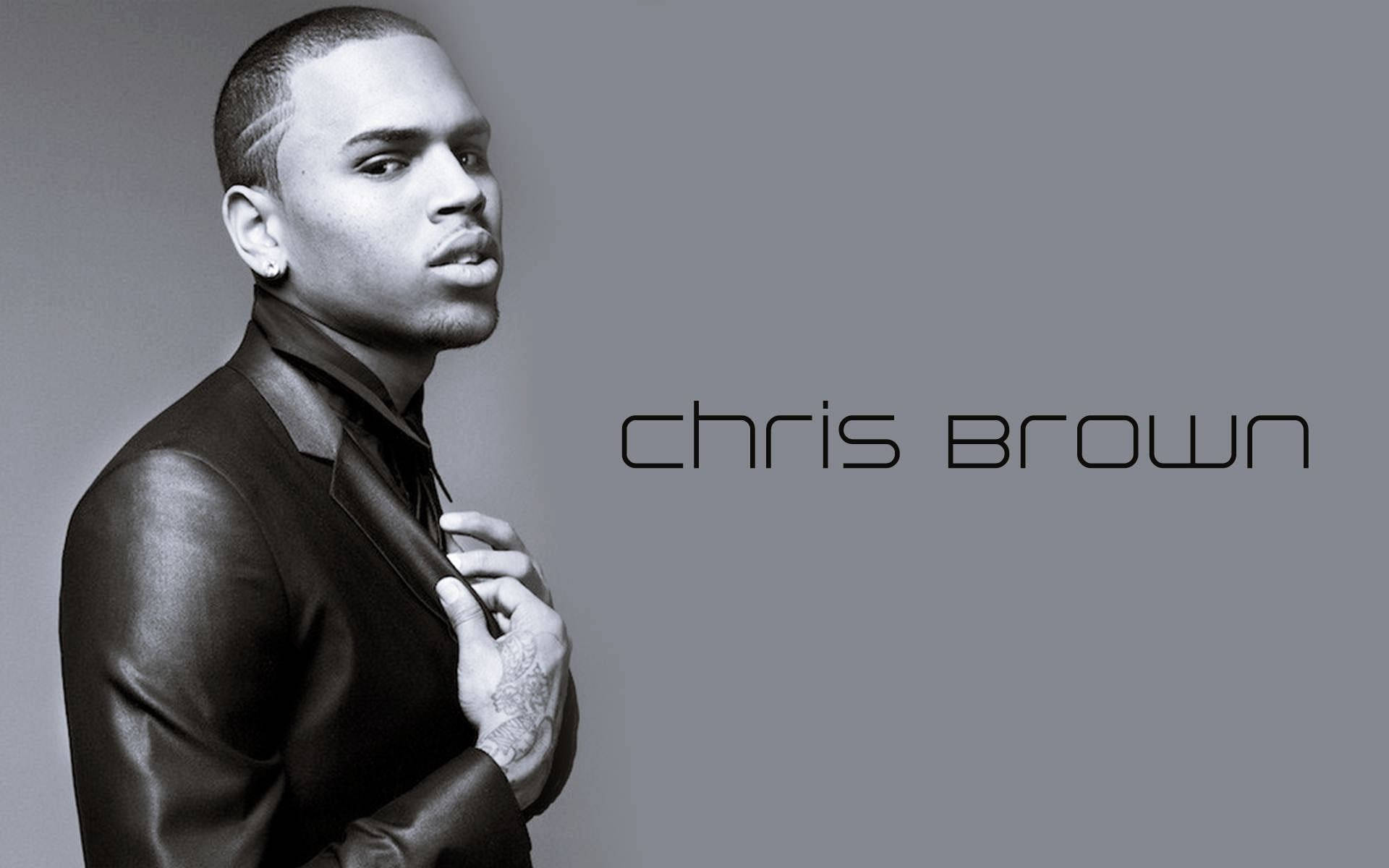 灰階克里斯·布朗穿西裝 壁紙 — Chris Brown身穿時尚黑色皮褸，在簡約灰色背景前自信擺姿勢，他的名字優雅地寫在旁邊。