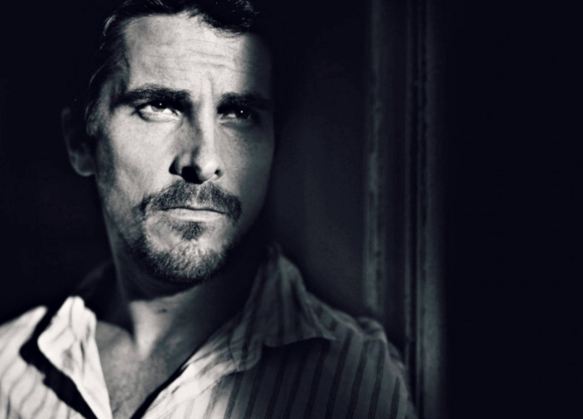 灰階克里斯蒂安·貝爾男性面部 壁紙 — 一個眼神銳利、鬍渣未刮的男人靠在黑暗門口，充滿情緒的黑白肖像。