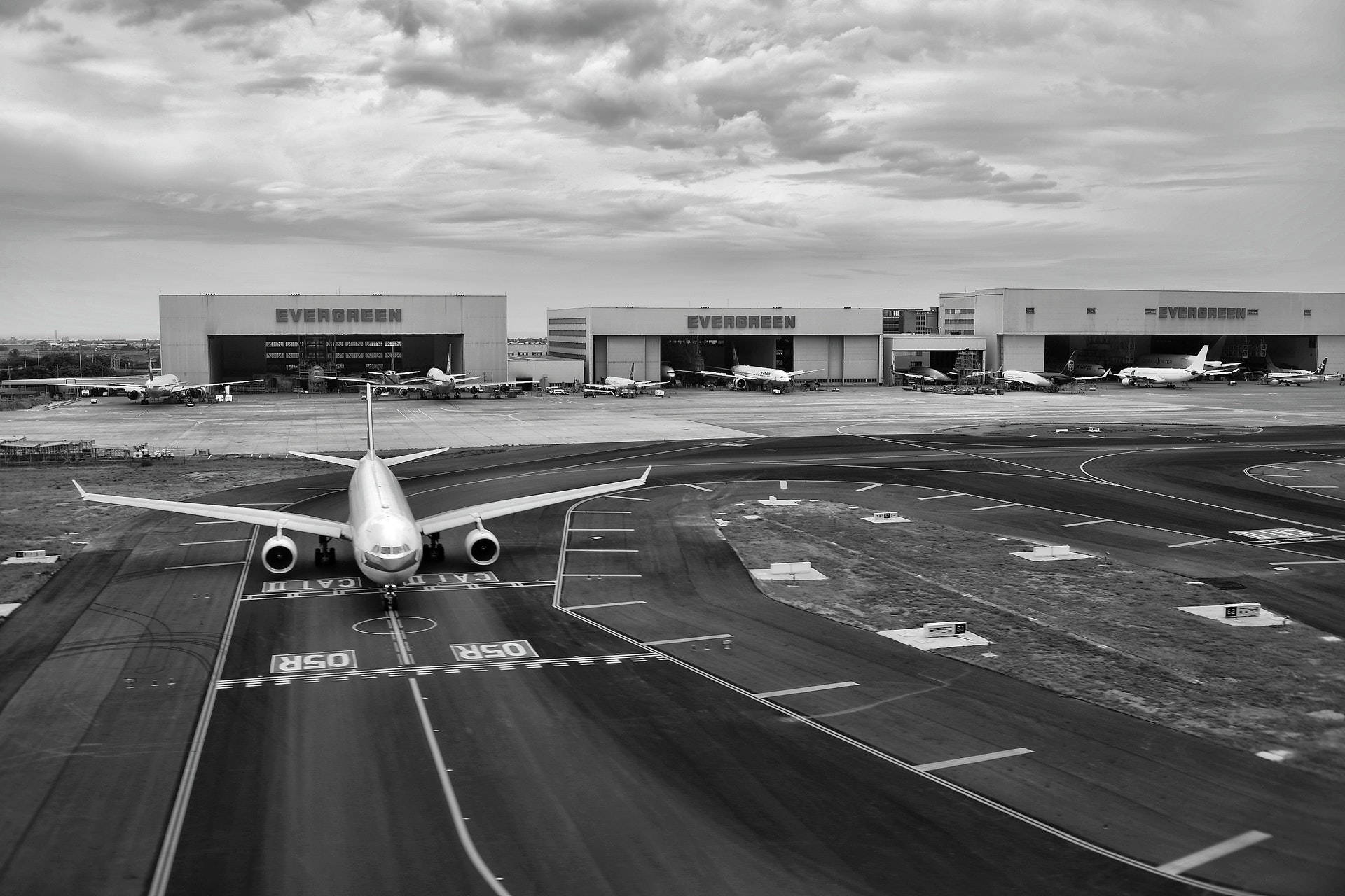 灰階機場跑道 壁紙 — 一架大型客機在機場停機坪上的黑白航拍圖，背景有機庫和其他飛機，天空多雲。