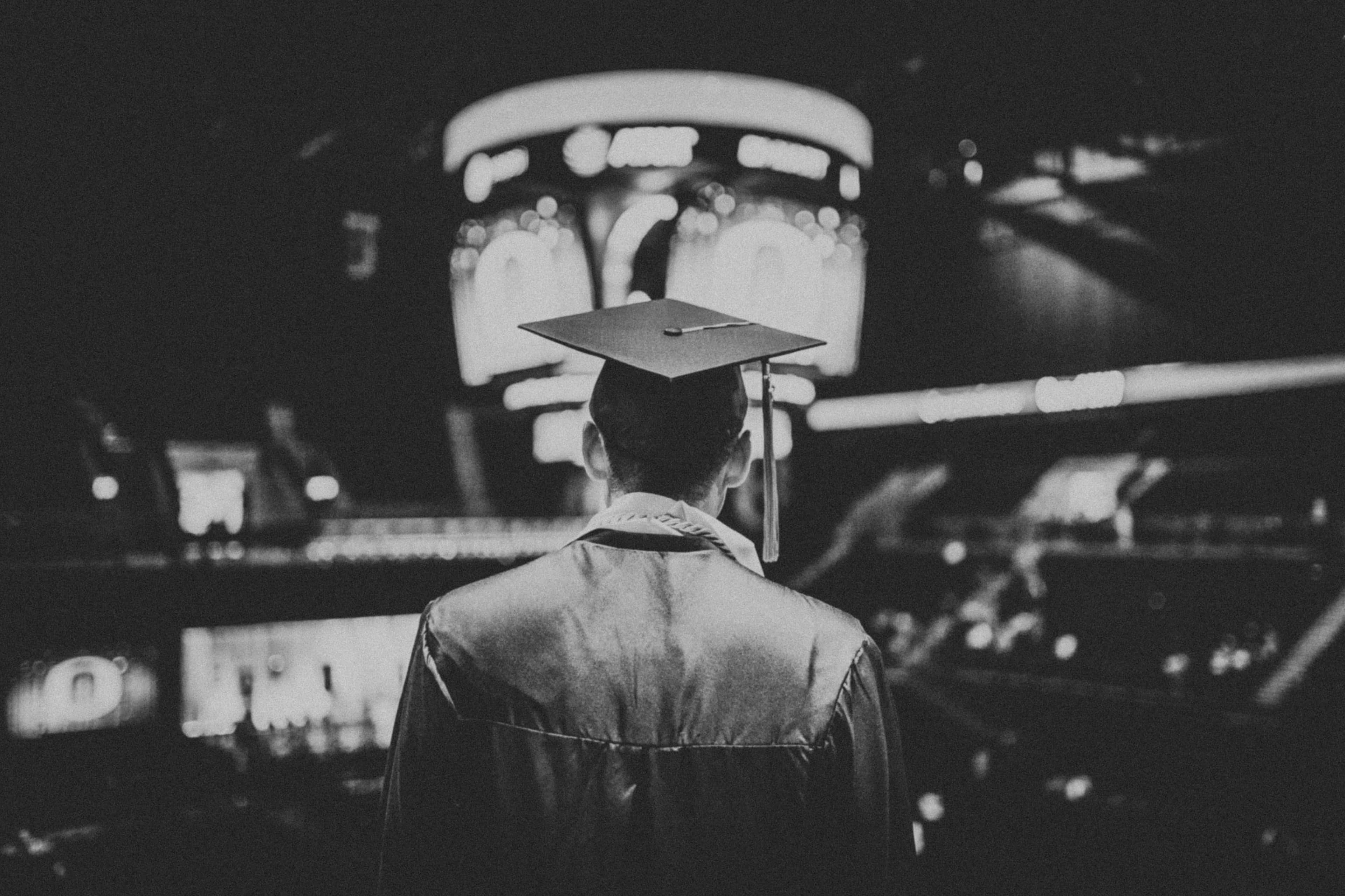 灰階男子戴畢業帽與學士服圖 壁紙 — 一位身穿畢業袍和四方帽的畢業生夜晚獨自站在體育館，凝視著明亮的記分牌。