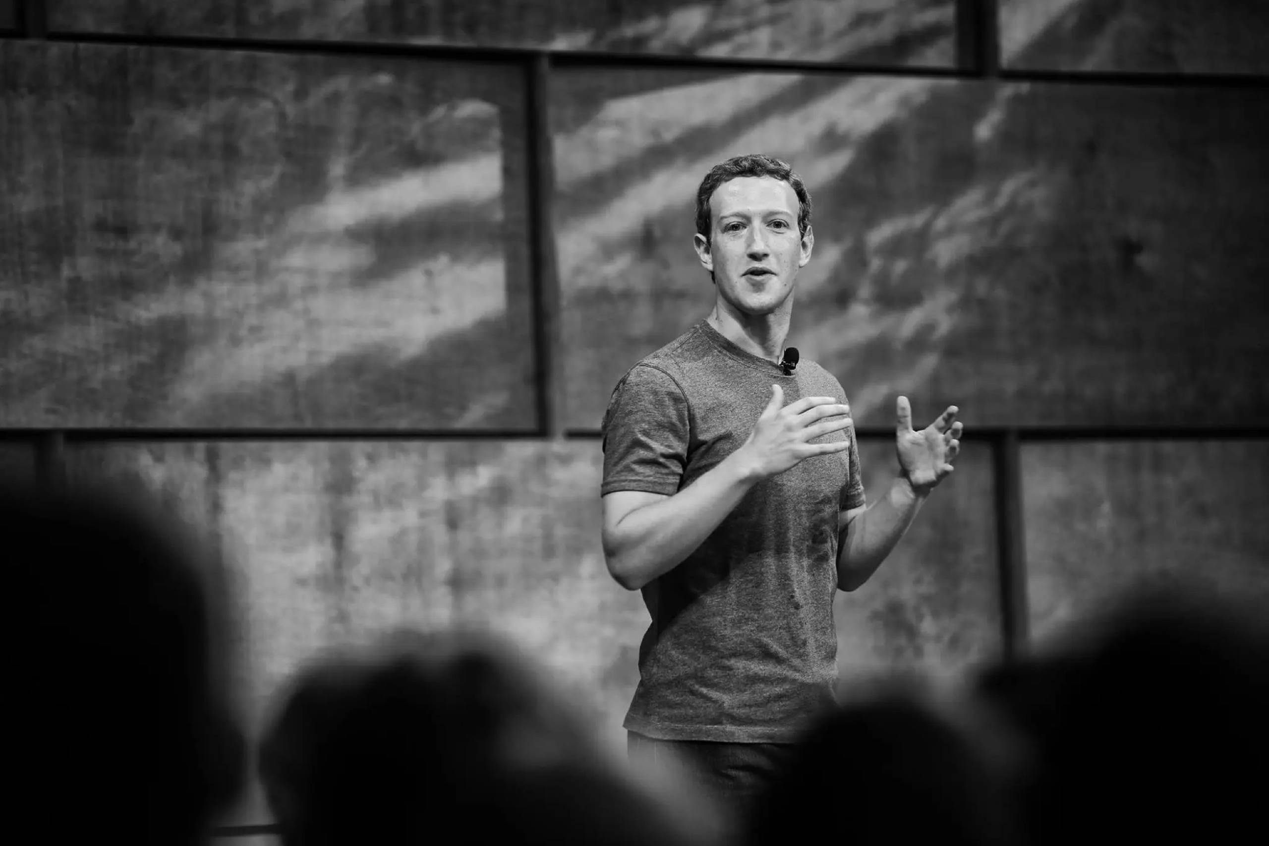 灰階馬克·祖克柏 壁紙 — 一名身穿T恤的男子在舞台上熱情演講，模糊的觀眾剪影以黑白呈現。