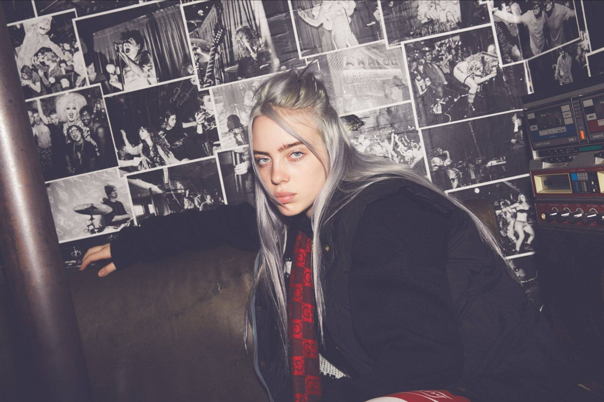灰髮比莉·艾利什 4K 壁紙 — Billie Eilish 喺昏暗嘅房間入面，靠喺一幅鋪滿黑白演唱會相片嘅牆上。