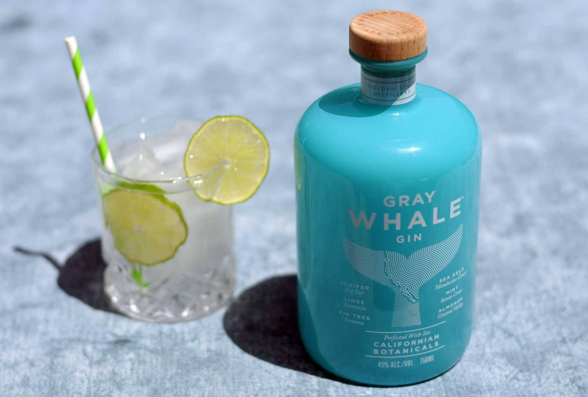 灰鯨琴酒檸檬杯 壁紙 — 在明亮的陽光下，一個藍綠色的Gray Whale Gin酒瓶和一個以青檸裝飾的氈酒雞尾酒放在有紋理的藍色表面上。