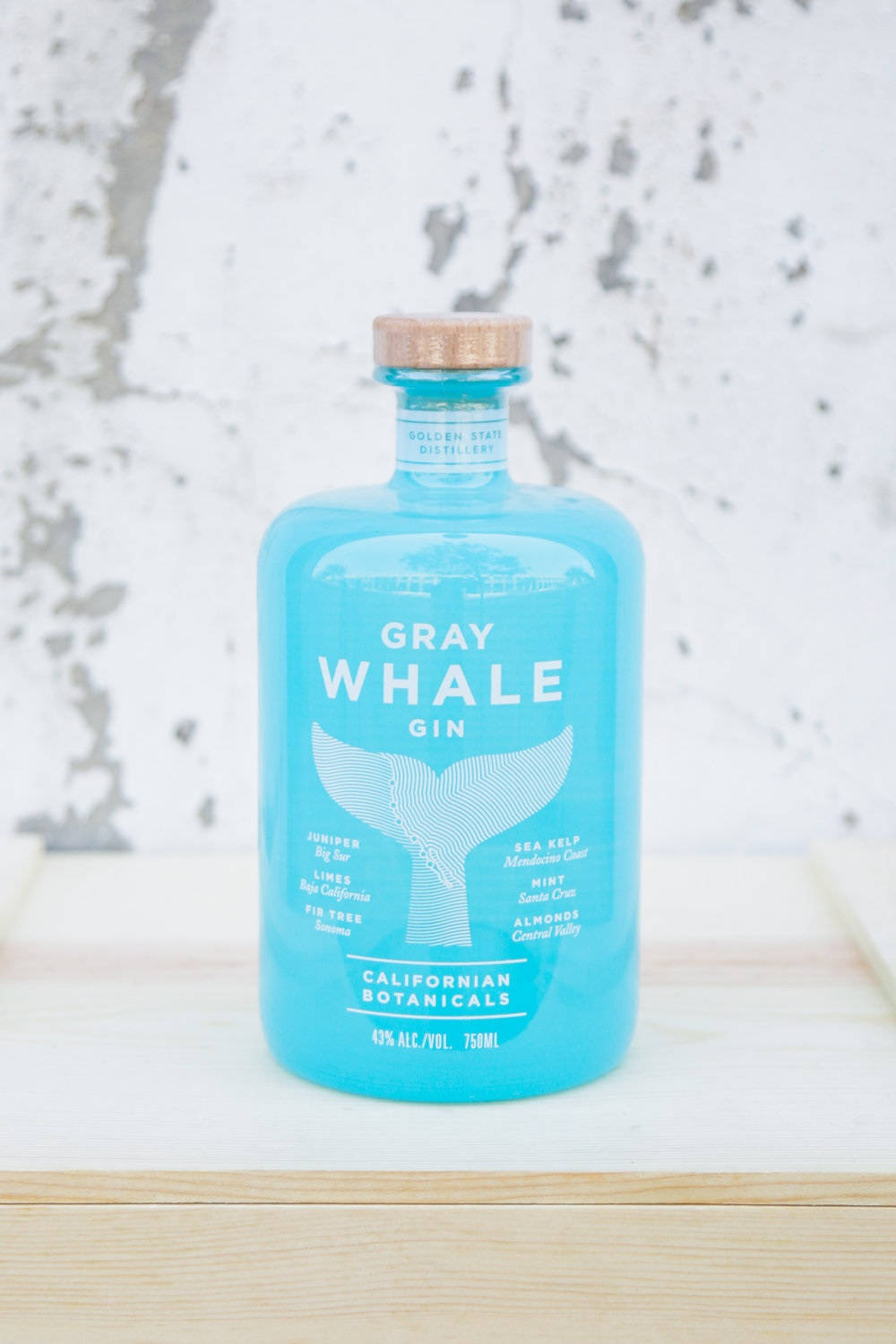 灰鯨琴酒白色 壁紙 — 一個充滿活力的藍綠色Gray Whale Gin酒瓶，帶有木質瓶蓋，置於淺色木質表面中央，背景為有紋理的白色牆壁。