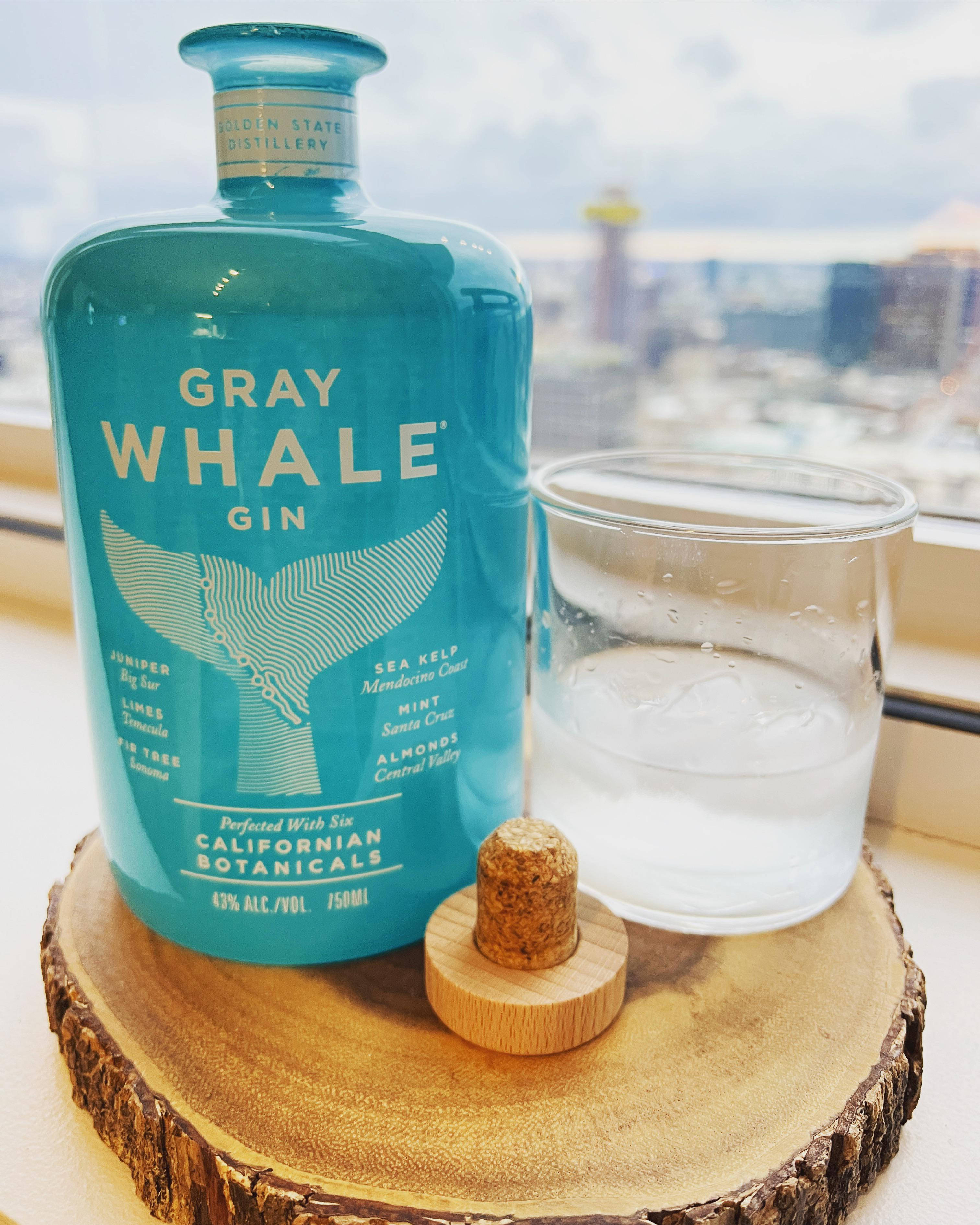 灰鯨琴酒窗邊 壁紙 — 一個藍綠色的Gray Whale Gin酒瓶放在木切片上，旁邊有一杯冰水，軟木塞放在杯墊上，透過窗戶俯瞰城市景觀。