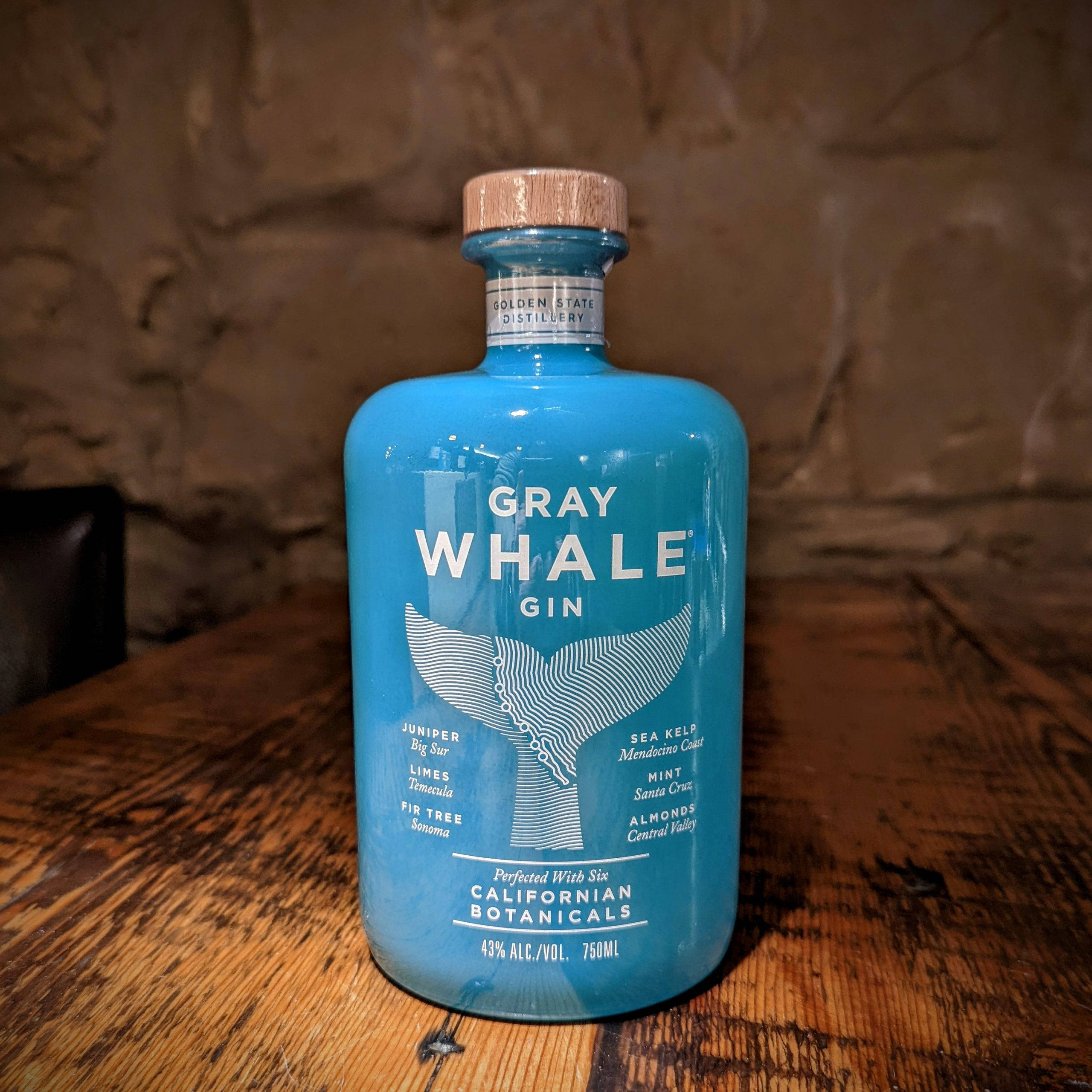 灰鯨琴酒肖像 壁紙 — 一個充滿活力的藍色Gray Whale Gin酒瓶，帶有鯨魚尾巴設計，放在一個質樸的木桌上，背景是石牆。