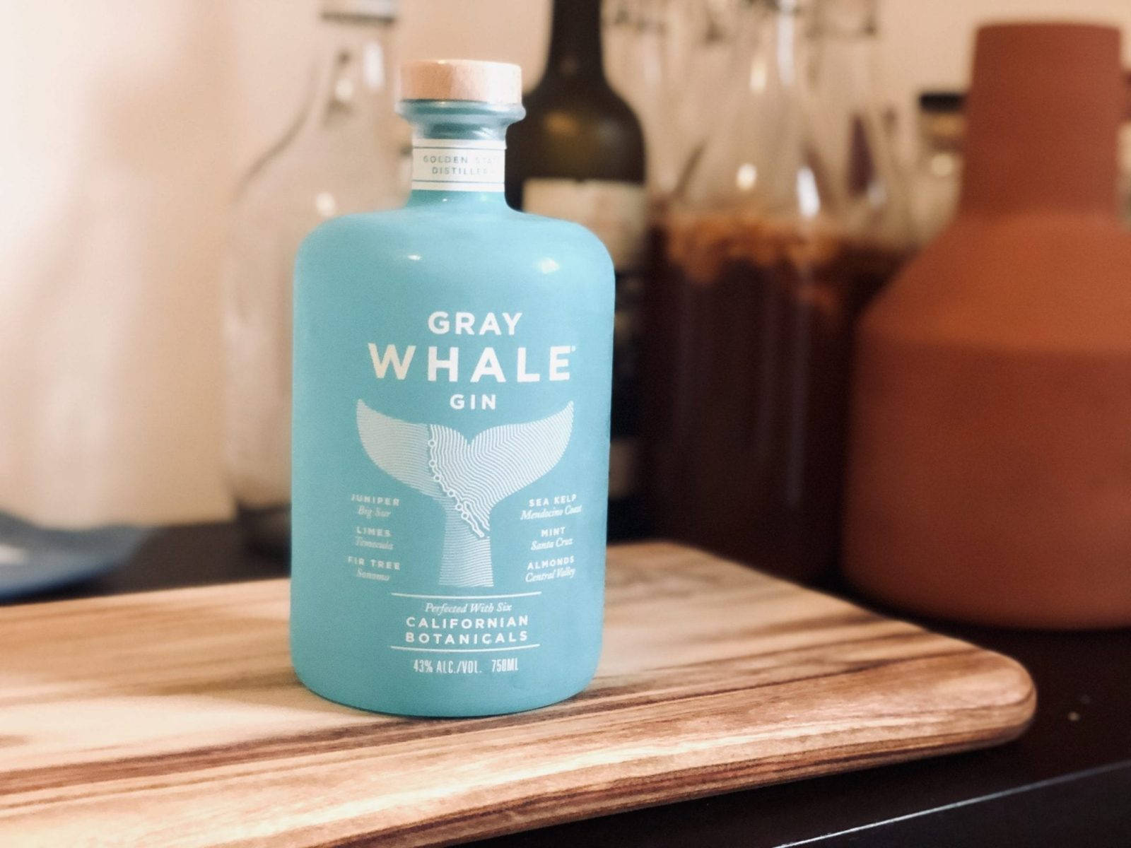 灰鯨金酒桌上擺設 壁紙 — 一個藍綠色的Gray Whale Gin酒瓶放在木製砧板中央，背景有模糊的廚房用具。