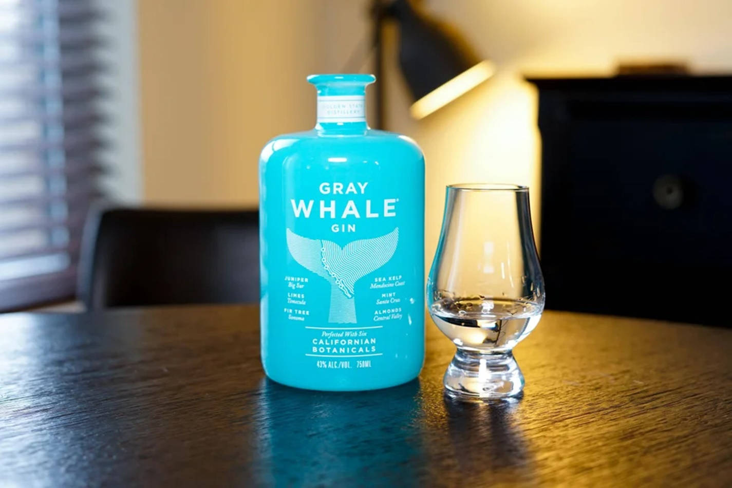 灰鯨金酒玻璃杯 壁紙 — 一瓶充滿活力的藍綠色Gray Whale Gin酒瓶放在一張溫暖的木桌上，旁邊有一個空的品酒杯，房間光線柔和。