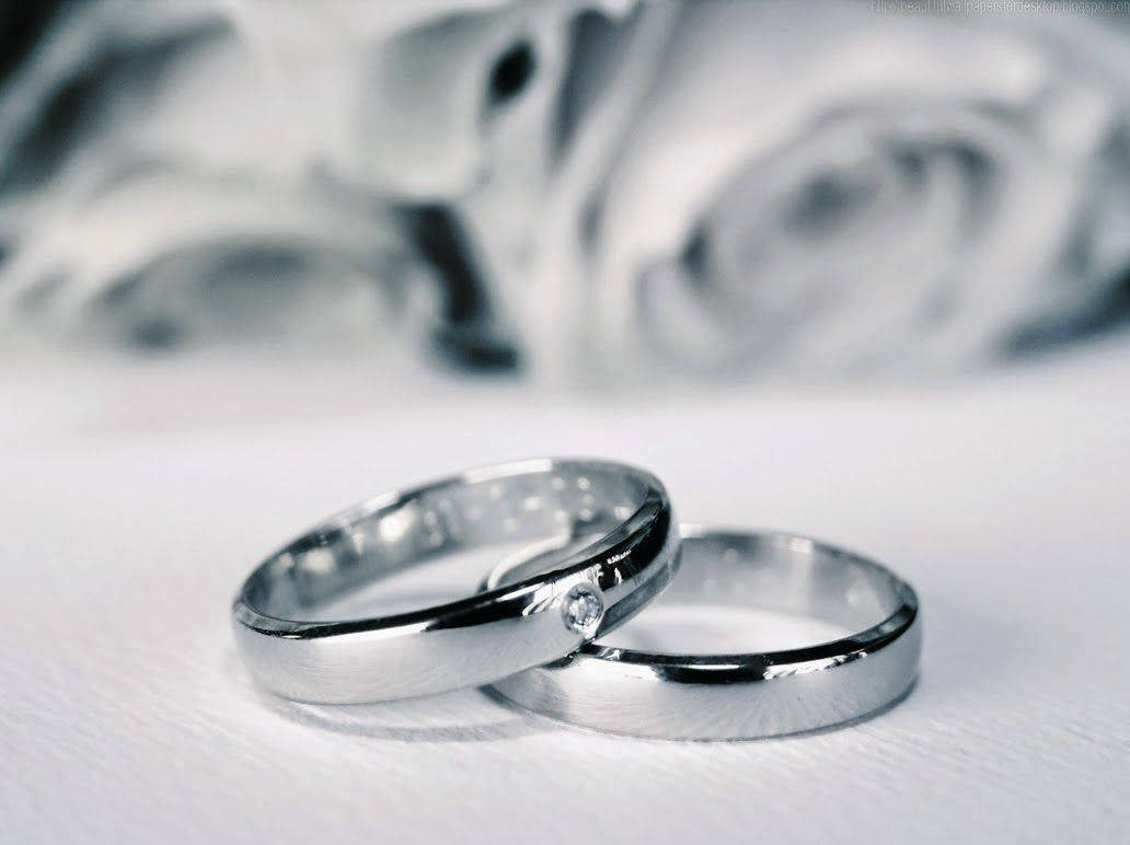 灰黑色婚戒 壁紙 — 兩枚銀色結婚戒指，鑲有一顆小鑽石，放在白色表面上，背景有模糊的玫瑰。