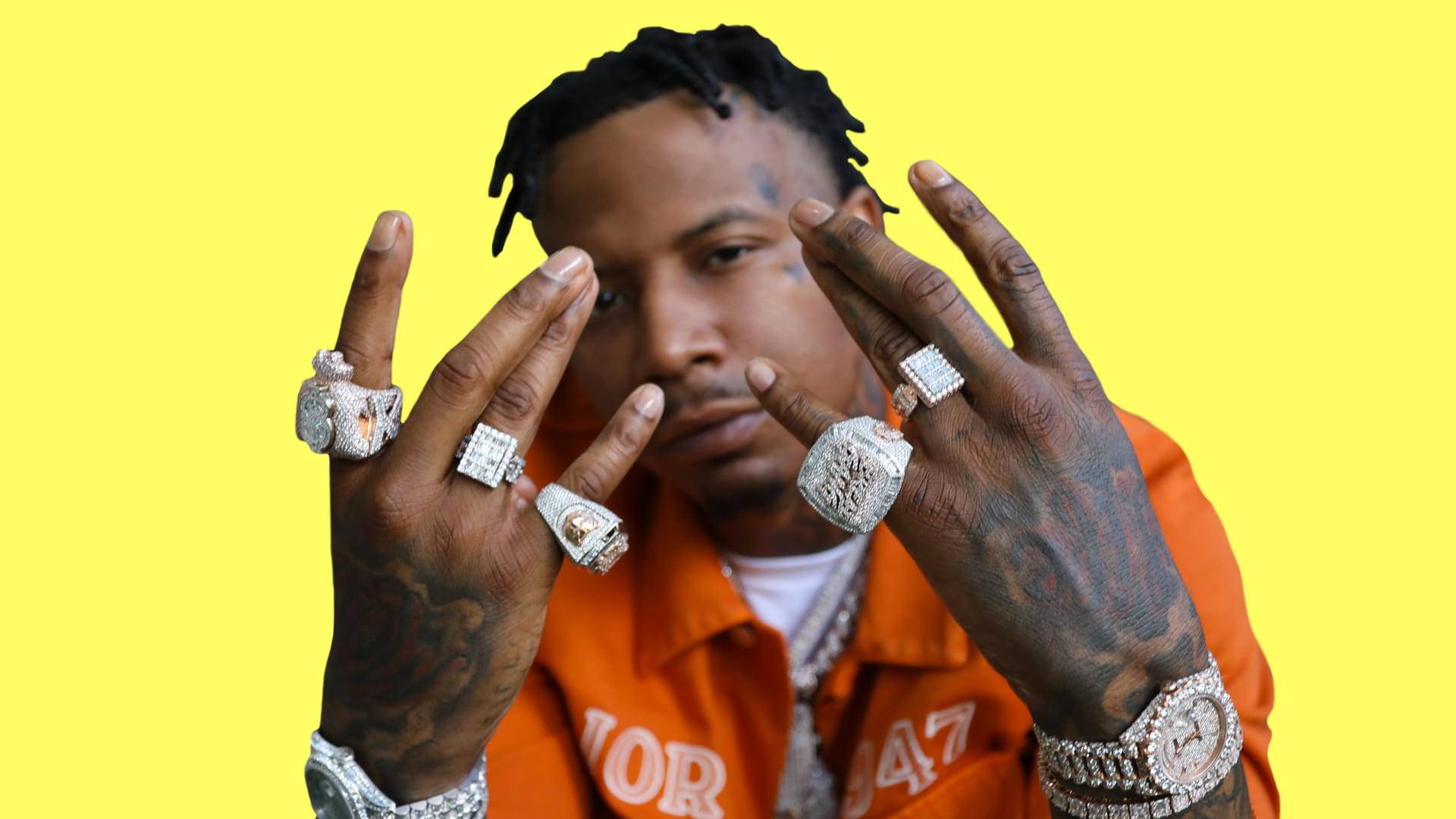 炫富 Moneybagg Yo 壁紙 — 一名留著辮子頭、身上有紋身的男子，在明亮的黃色背景前佩戴多枚鑽石戒指和手錶。