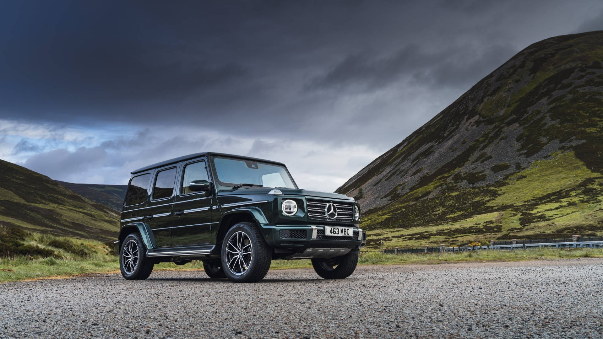深綠色平治 G-Class SUV 停泊在礫石路上，背景有山脈和暴風雨的天空。