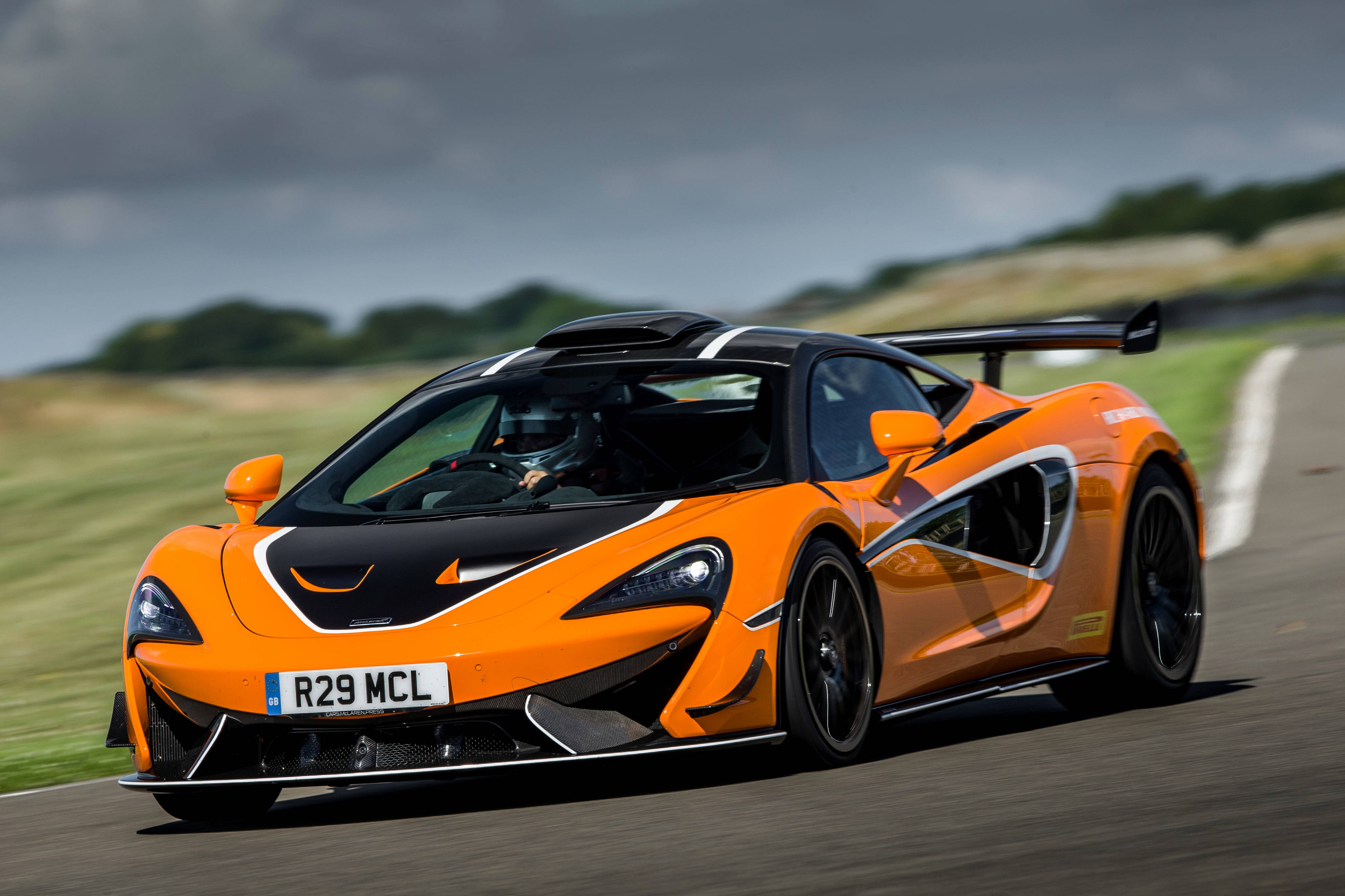 炫目橙色賽車動態圖 壁紙 — 一輛充滿活力的橙色McLaren跑車在賽道上疾馳，背景是模糊的綠色山丘和陰天。