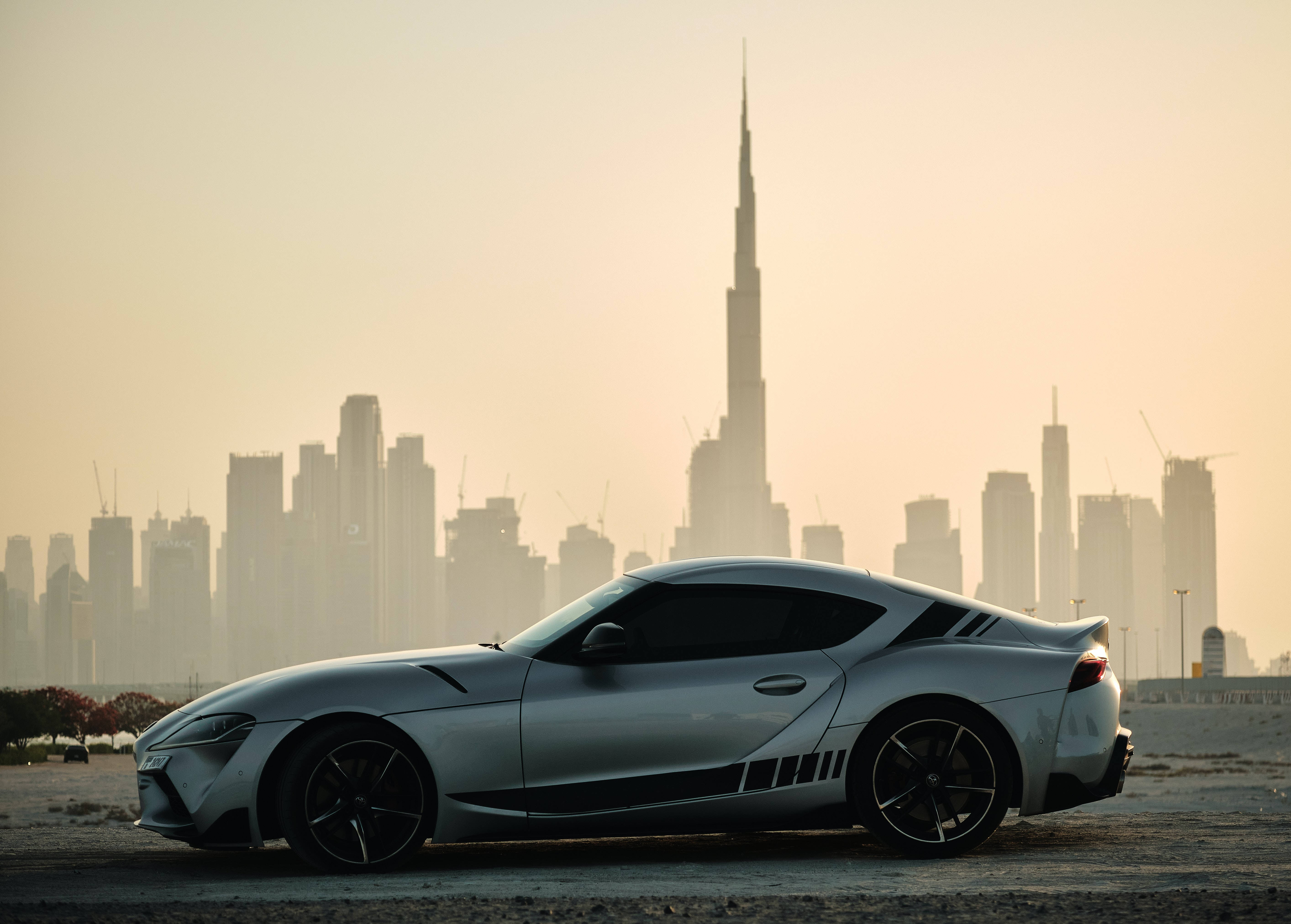 炫酷 4K 《Toyota Supra》灰銀色搭配黑色細節 壁紙 — 一架時尚的銀色跑車停在沙漠地形上，杜拜的哈利法塔和天際線在金色時刻的薄霧中閃耀。