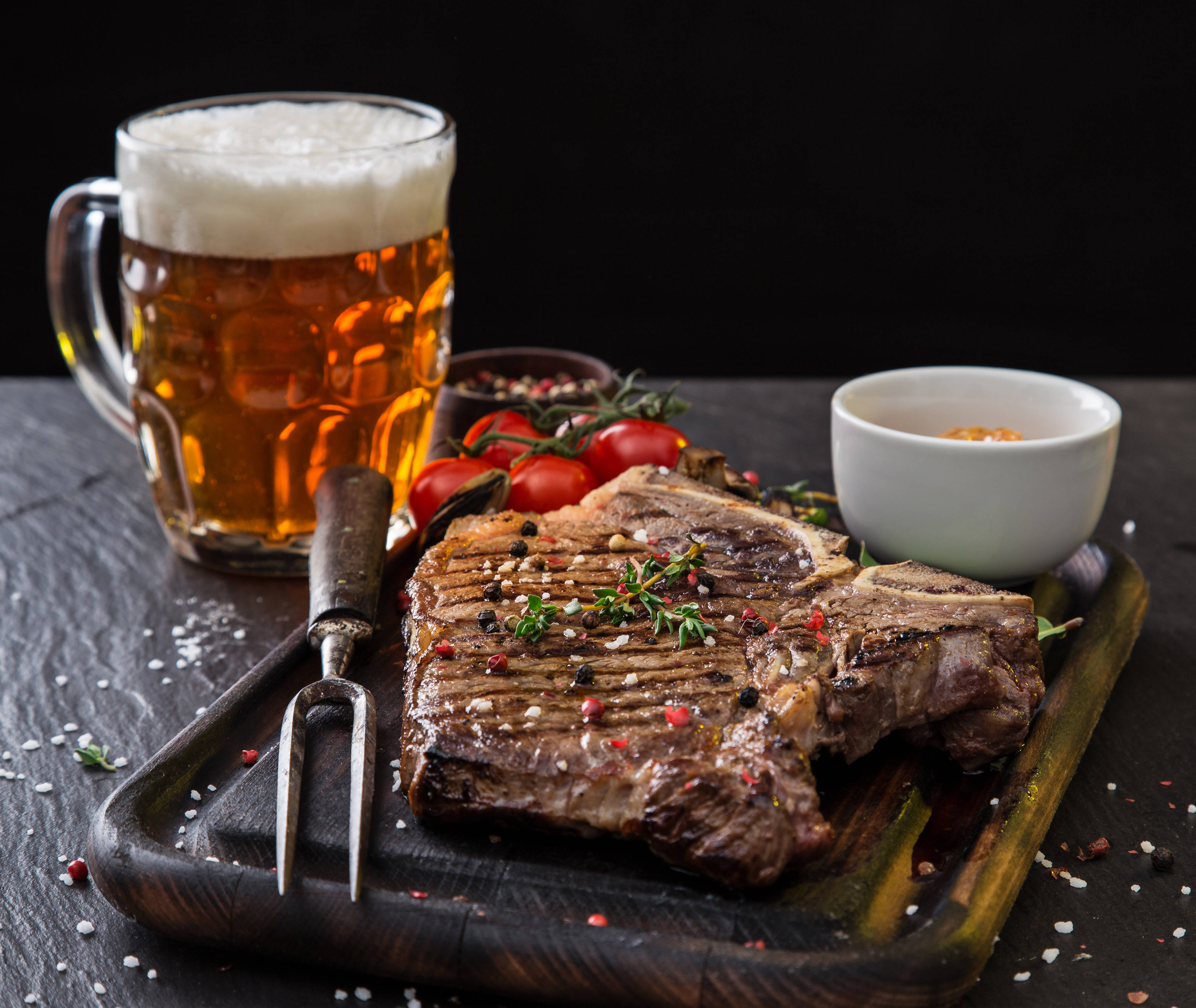 炭烤肉配啤酒 壁紙 — 一塊多汁的烤T骨牛扒，用香草和香料調味，放在木板上，旁邊有一杯泡沫啤酒和車厘茄。