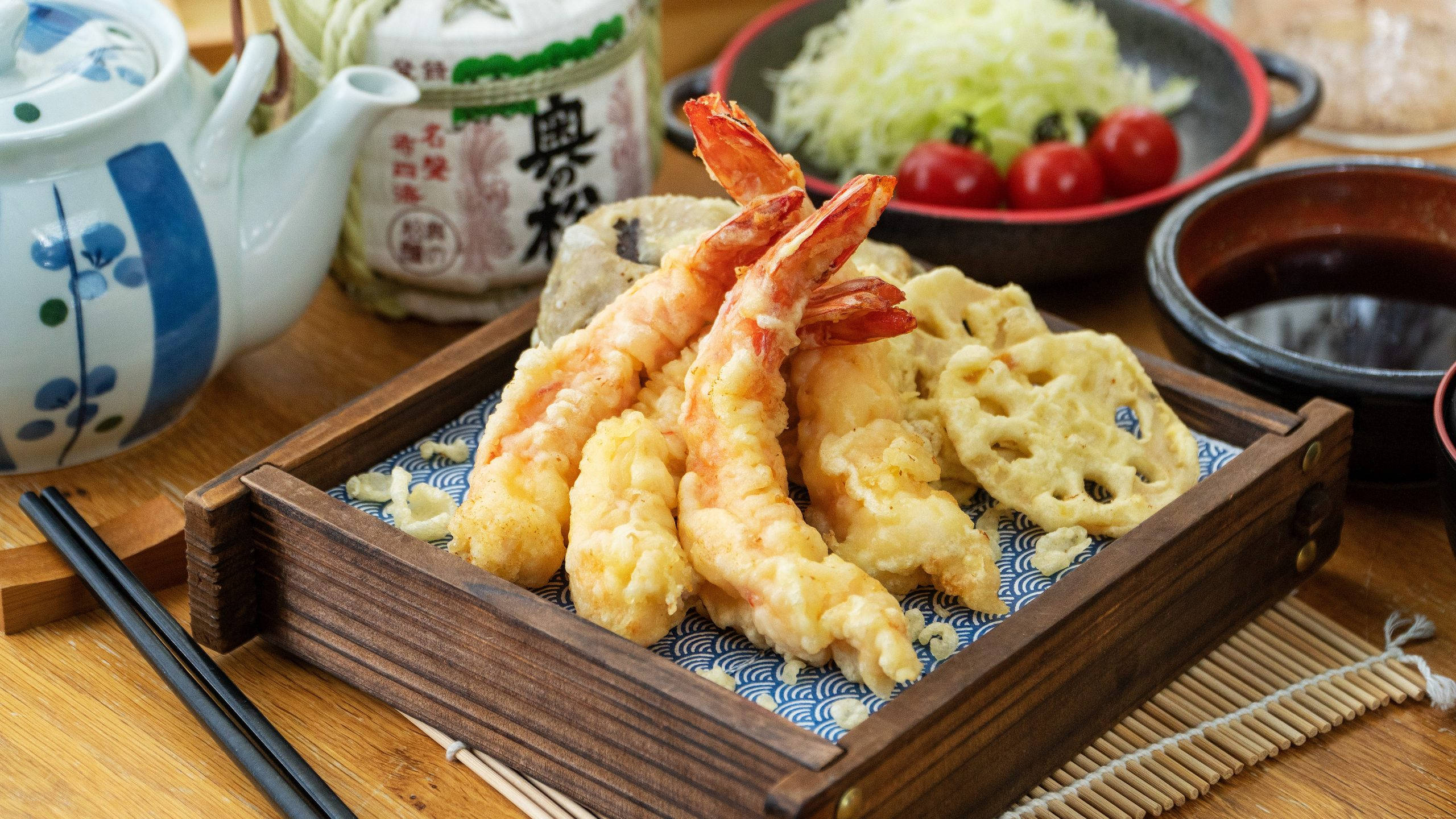 炸天婦羅與番茄 壁紙 — 金黃色天婦羅蝦和蓮藕，盛載於木盒中，配以小菜和茶，置於竹蓆上。