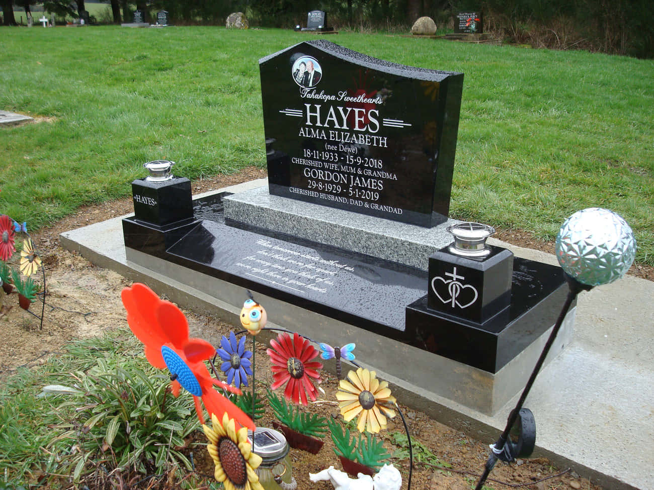 Memorial 桌布 壁紙 — 為Alma Elizabeth Hayes和Gordon James Hayes打造的拋光黑色花崗岩墓碑，在草地墓園中以彩色金屬花朵和反光球體裝飾。