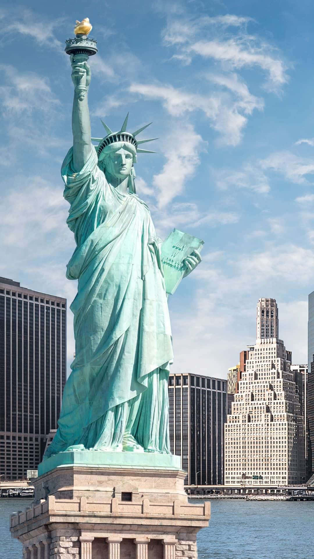 為你的手機裝點紐約城市美景 壁紙 — 自由女神像在藍天白雲下高高聳立，周圍是紐約市的高樓大廈和水域。