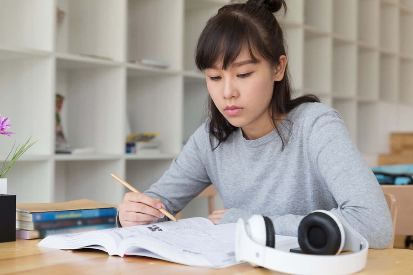 為夢想努力奮鬥 壁紙 — 一位專注的年輕女性穿著灰色毛衣，在木桌上寫筆記，附近有耳機，背景有紫色花朵。